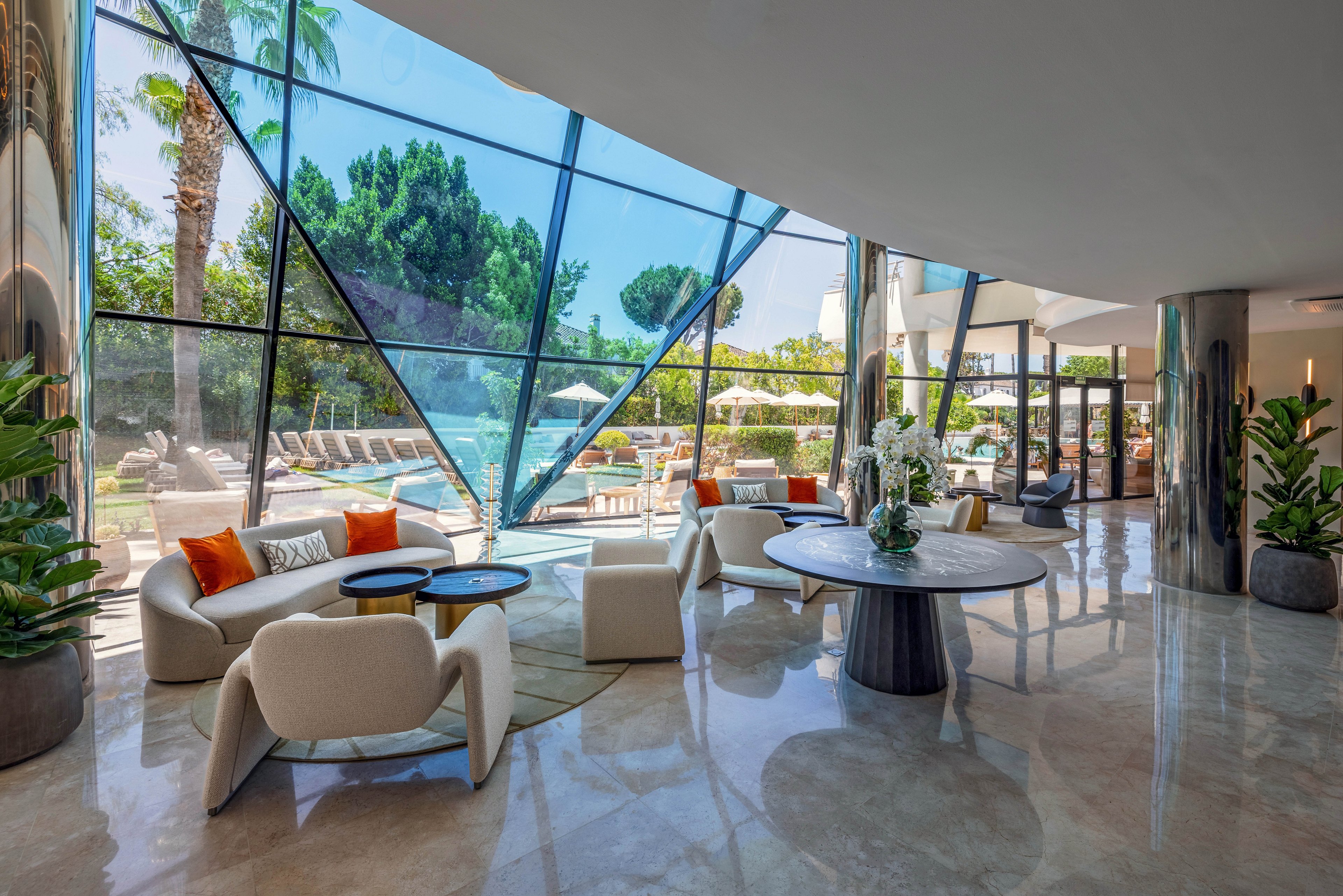 Contemporary lounge seating is arranged in a spacious hotel lobby with large floor-to-ceiling glass windows. The interior features curved sofas, armchairs, and round tables on a polished stone floor. Outside, sun loungers and umbrellas surround a swimming pool framed by lush greenery and palm trees. Natural light floods the space, highlighting the clean, modern design and relaxed resort atmosphere.