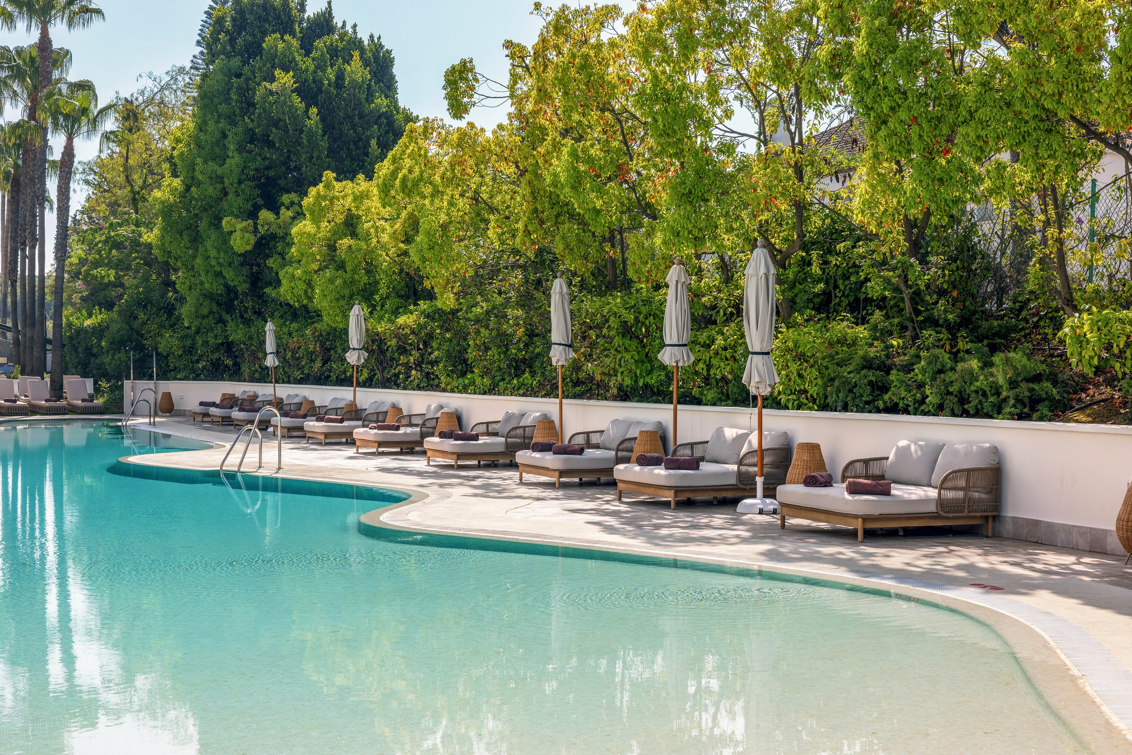 An inviting outdoor swimming pool is surrounded by cushioned sun loungers and closed umbrellas. The scene is set in a resort-style garden with lush green trees and palms behind a white wall. Clear turquoise water and a shallow entry create a relaxing atmosphere. The overall mood is tranquil and summery, suggesting a peaceful vacation setting.