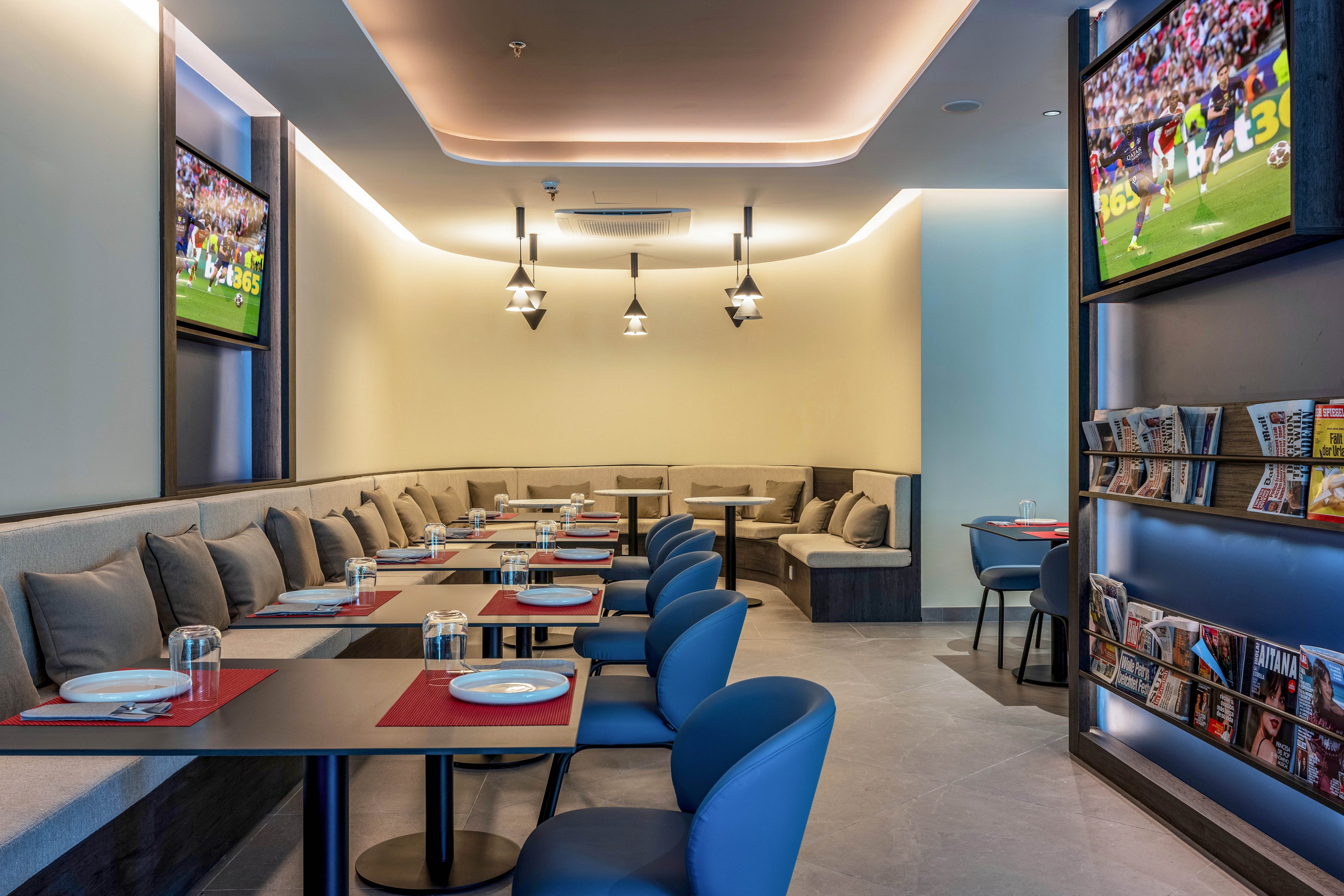 Contemporary restaurant interior features neatly arranged tables with red placemats and white plates along a cushioned banquette. Multiple flat-screen televisions on the walls display a sports game, creating a casual lounge atmosphere. Soft ceiling lighting and pendant lamps highlight the clean, modern design. A magazine rack filled with reading materials adds to the relaxed, entertainment-focused setting.