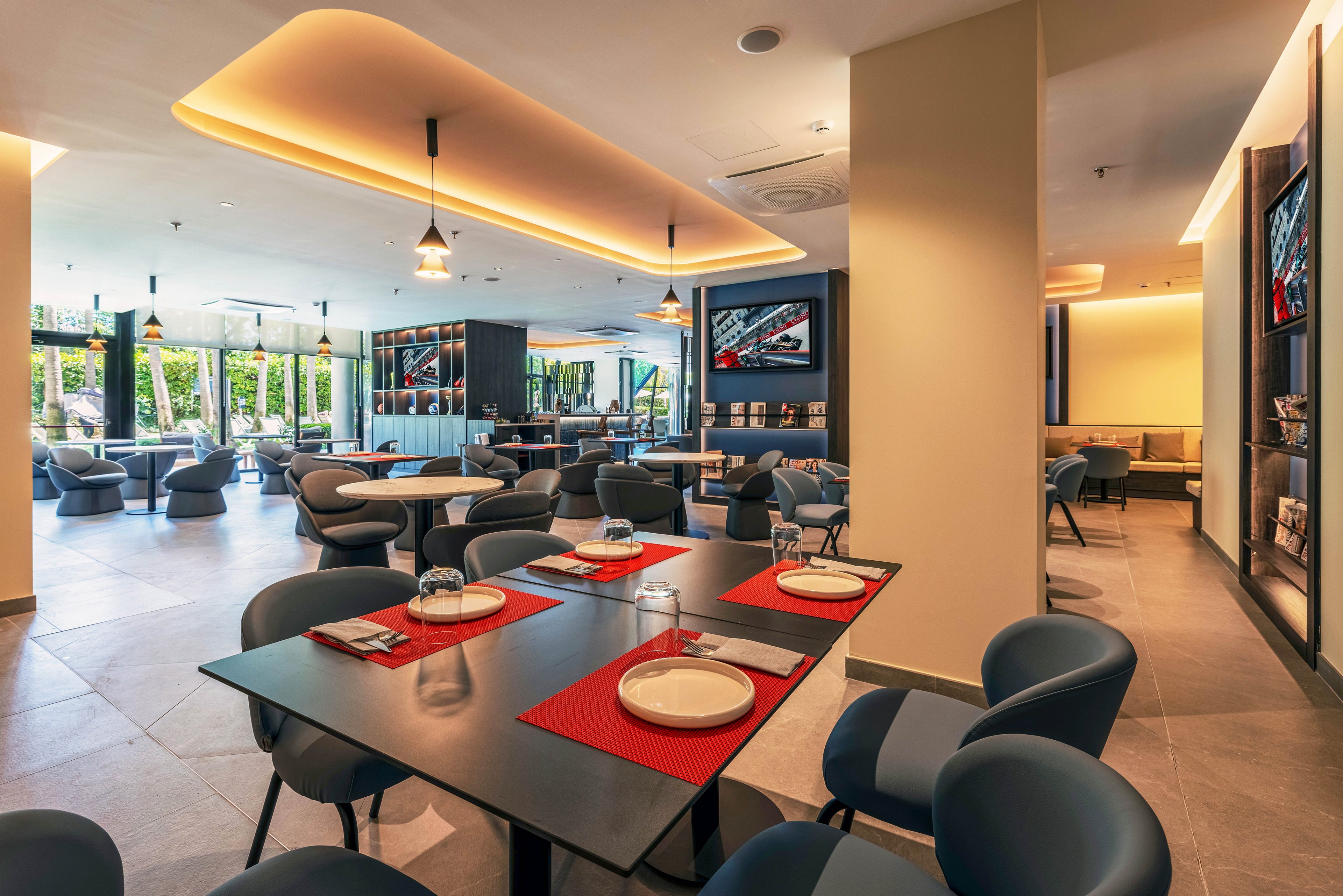 Contemporary restaurant lounge interior featuring dark tables set with white plates and red placemats. Comfortable armchairs and low seating areas extend toward large floor-to-ceiling windows with garden views. Warm ceiling lighting and pendant lamps create a cozy atmosphere, while wall-mounted screens and shelving add a stylish, upscale touch. The space appears clean, spacious, and designed for relaxed social dining.
