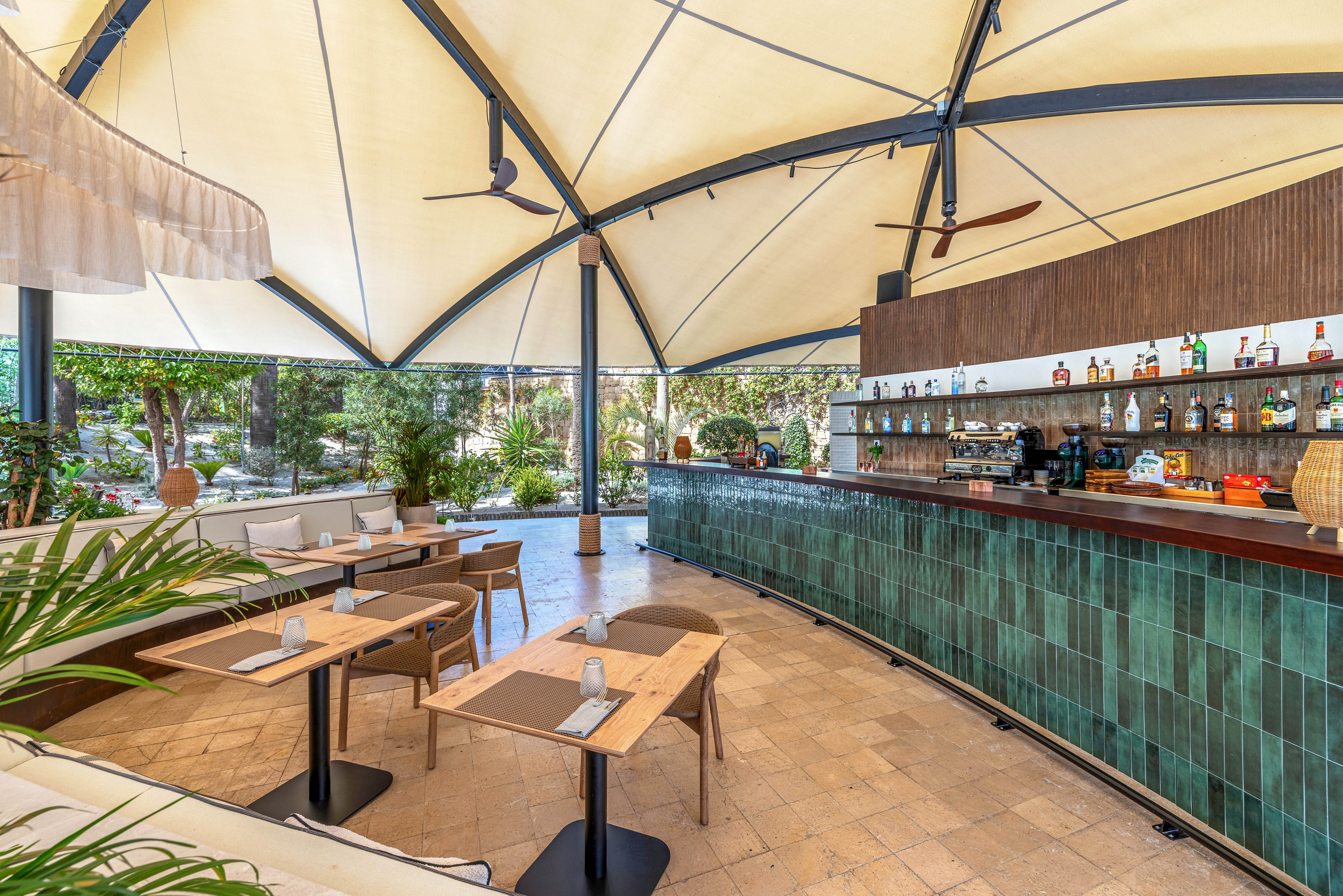 A contemporary open-air bar features a long counter clad in green tiles beneath a large white canopy roof. Wooden tables and chairs are neatly arranged on a stone floor, with napkins and glasses set for guests. Shelves behind the bar display various bottles and glassware, while plants and trees surround the seating area, creating a lush garden atmosphere. The space feels bright, relaxed, and inviting for casual dining or drinks.