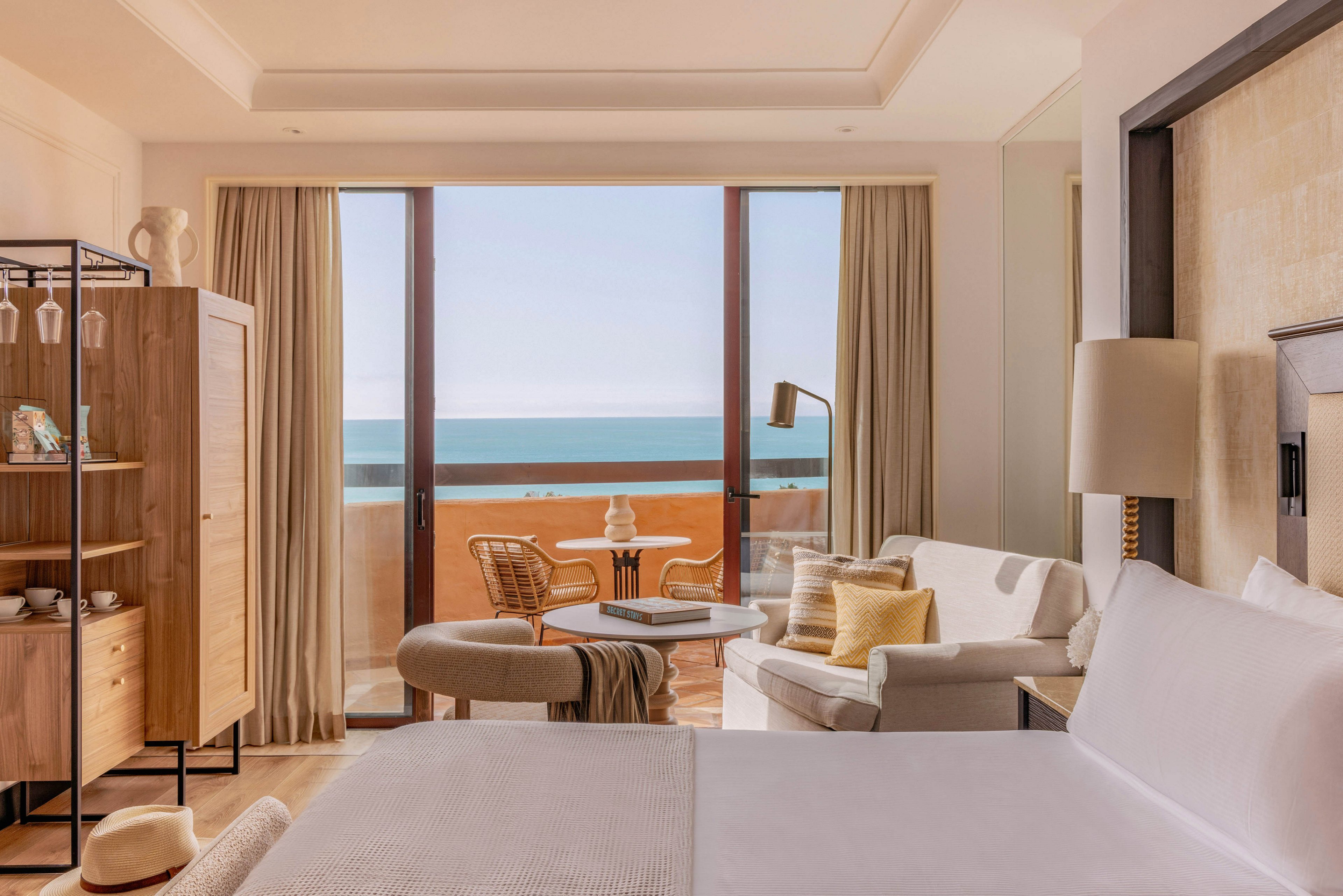 A sunlit hotel room features a neatly made bed in the foreground and a cozy seating area with armchairs and a small round table. Large floor-to-ceiling windows open onto a balcony overlooking a calm blue ocean. Neutral tones of beige, white, and light wood create a warm, minimalist aesthetic. Soft curtains, cushions, and a standing lamp add comfort and a relaxed vacation atmosphere.