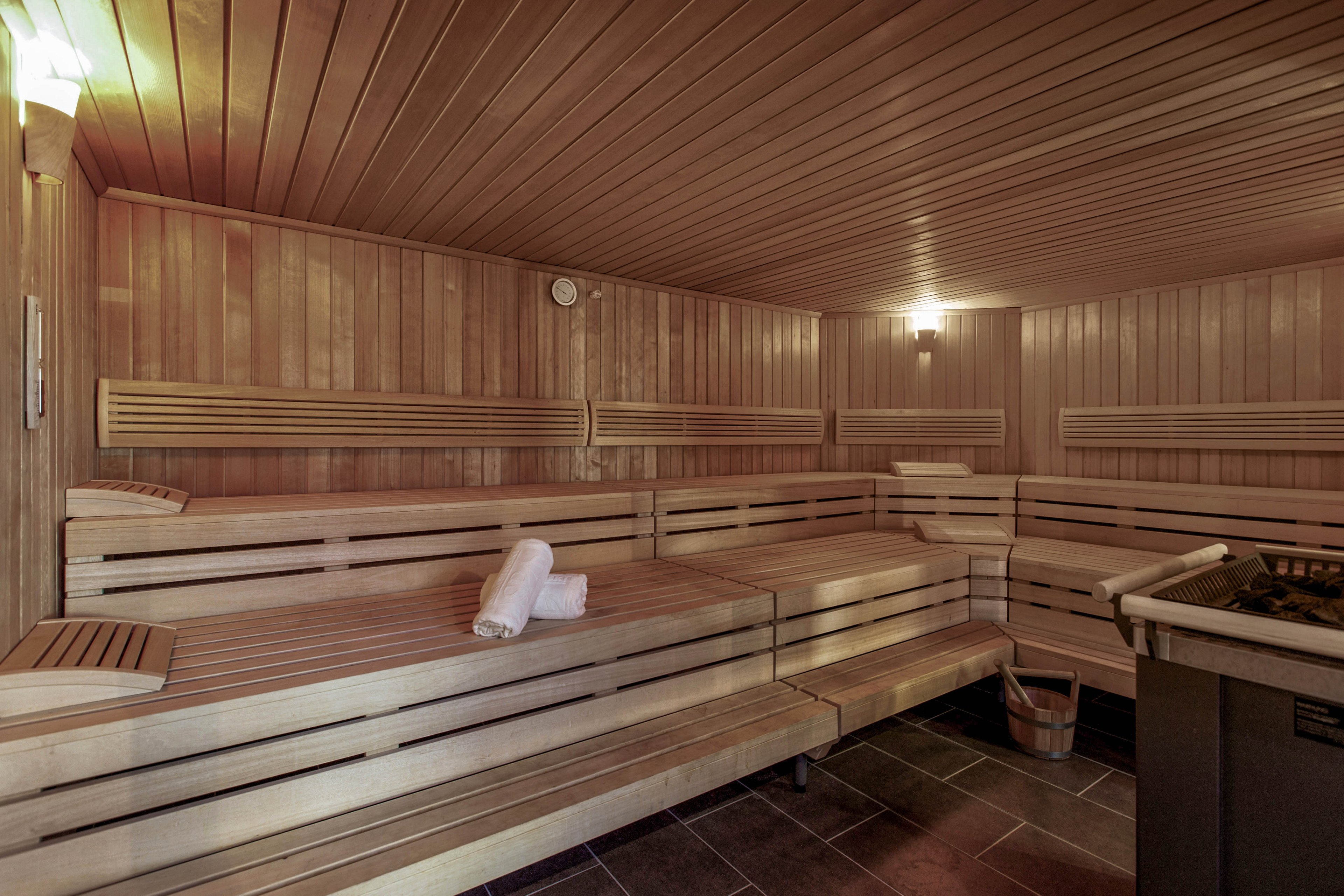 Spacious wooden sauna room with tiered benches is shown empty and ready for use. Rolled white towels rest on the middle bench, adding a touch of spa comfort. Warm ambient lighting highlights the wood paneling on the walls and ceiling. A sauna heater with stones is visible in the corner, emphasizing the relaxing wellness environment.