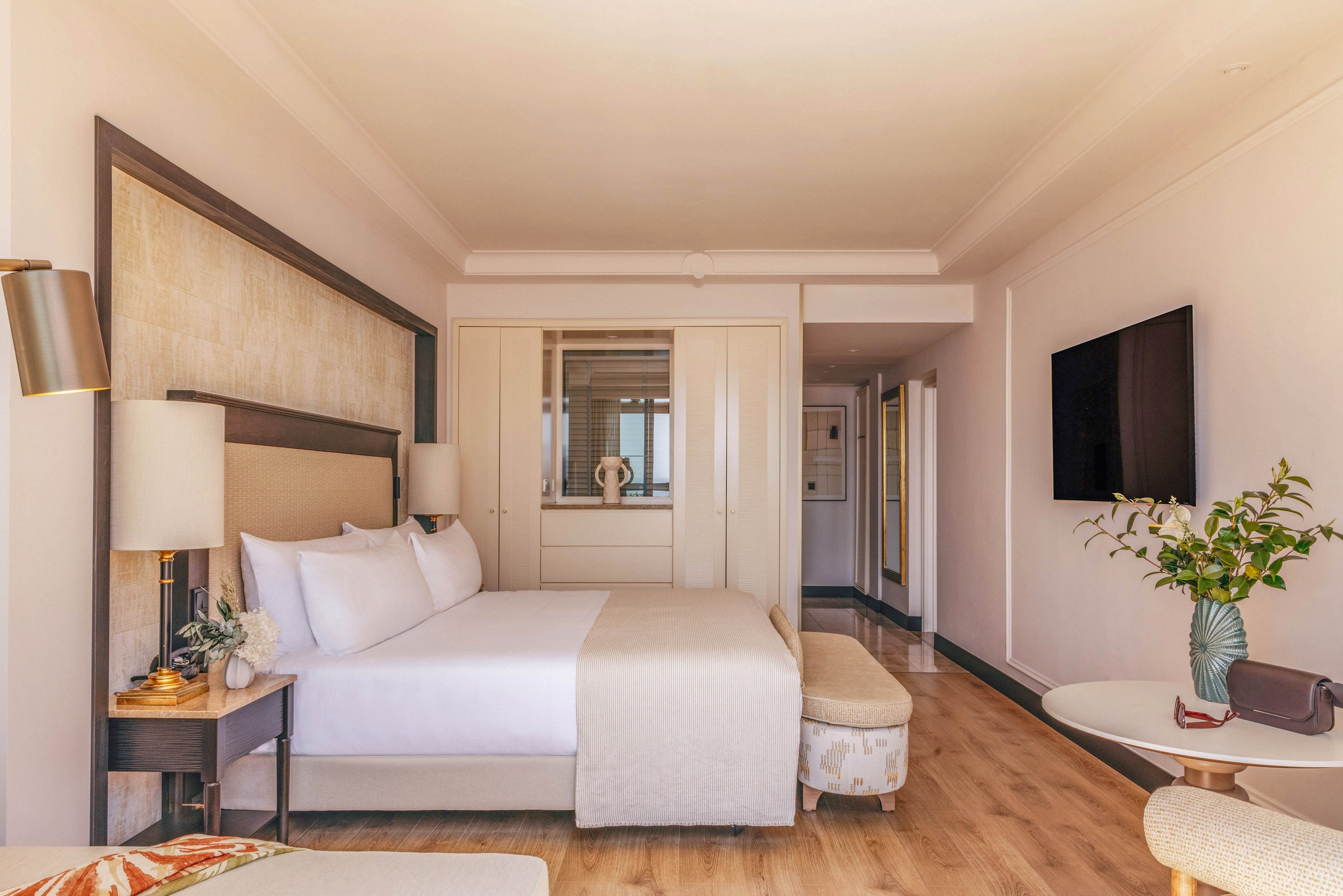 A spacious hotel bedroom features a large neatly made bed with white linens and a beige throw. The room includes a bedside table with a lamp, a cushioned bench at the foot of the bed, and a wall-mounted flat-screen television. Light wood flooring and soft neutral tones create a warm, inviting atmosphere. A small round table with a vase of green foliage and a handbag adds a touch of personal style to the contemporary interior.