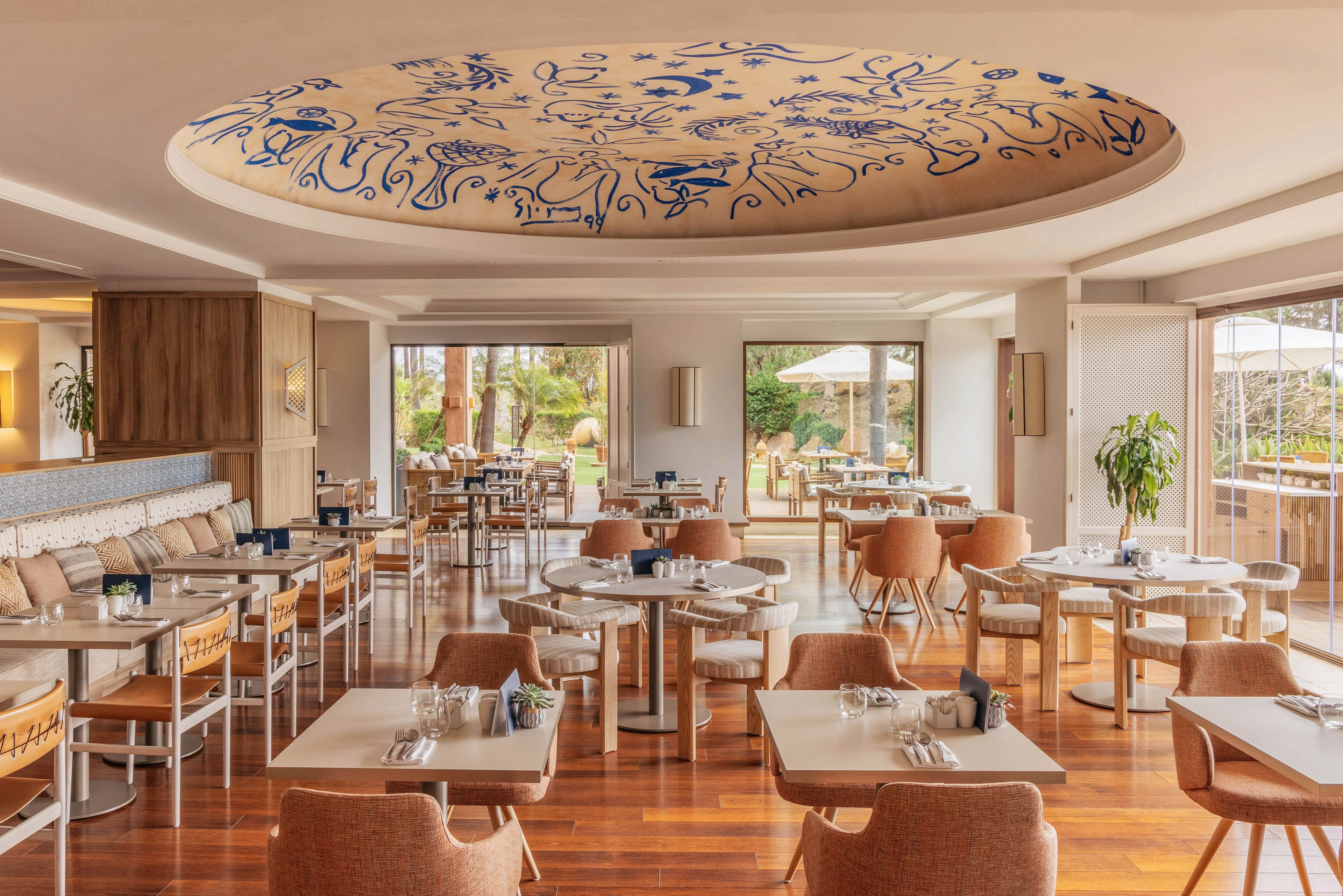 A spacious contemporary restaurant features neatly arranged tables and cushioned chairs on polished wooden floors. Large windows and sliding glass doors open to an outdoor terrace with greenery and umbrellas, filling the room with natural light. A circular recessed ceiling with blue abstract artwork serves as a striking focal point above the dining area. The warm neutral color palette and soft furnishings create an inviting, relaxed atmosphere suitable for casual or upscale dining.