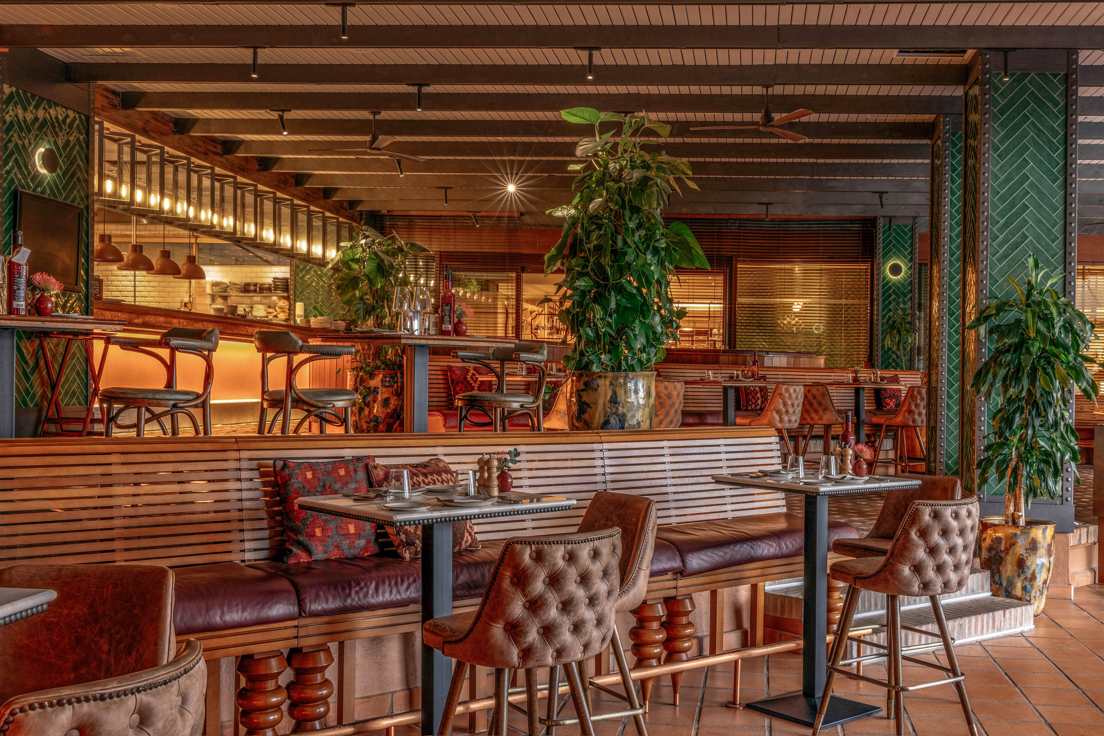 Warmly lit restaurant interior featuring leather chairs and wooden tables. The space includes a long bar counter with high stools and decorative lighting. Green plants and patterned cushions add texture and color throughout the room. The overall atmosphere feels inviting, contemporary, and comfortable for dining or socializing.