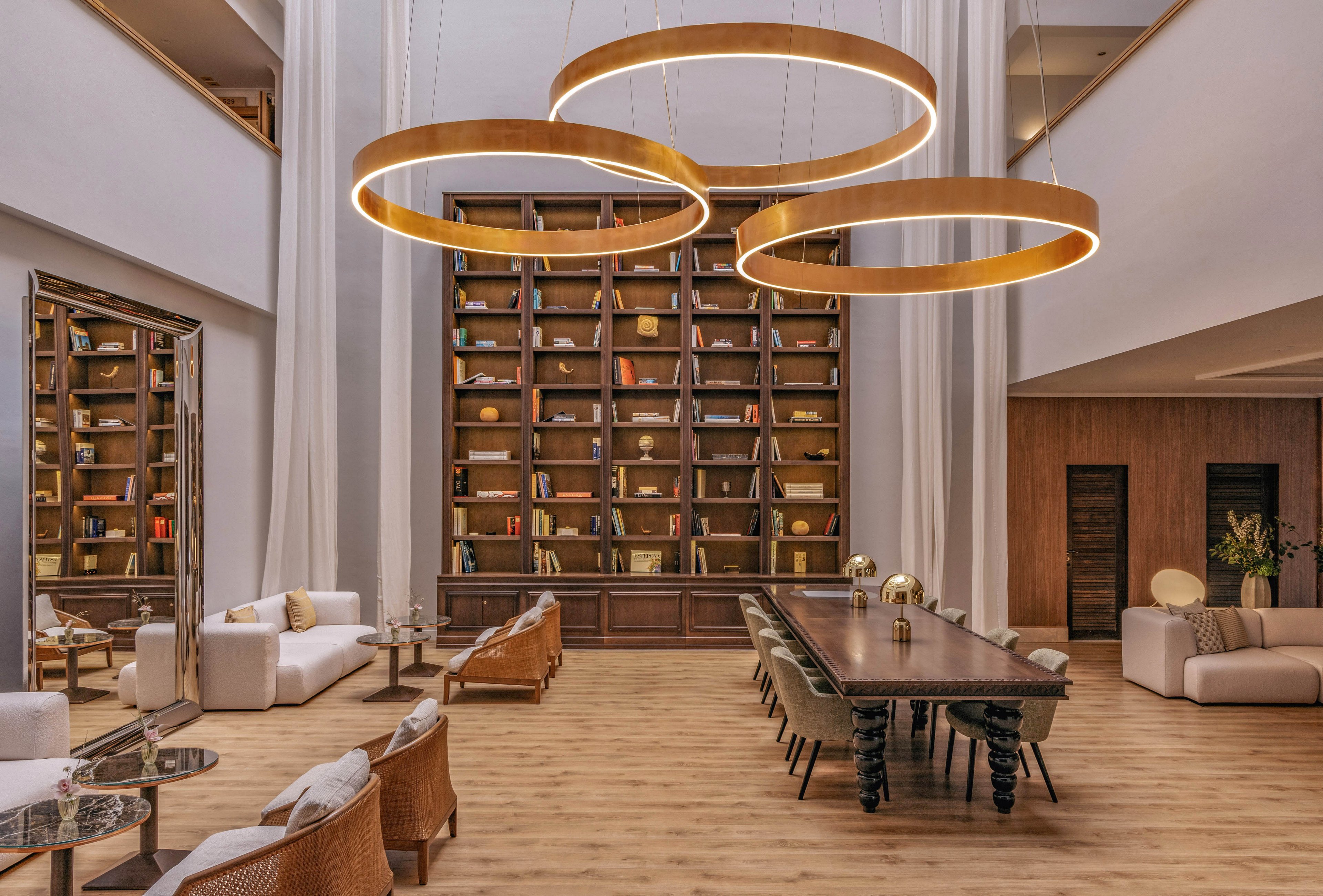 Spacious contemporary lobby features a towering built-in bookcase filled with books and decor. A long wooden communal table with dark legs sits centrally beneath three large circular pendant lights. Comfortable sofas and woven armchairs create multiple seating areas around low glass and marble tables. Warm wood flooring, high ceilings, and neutral furnishings give the space an upscale, inviting atmosphere.