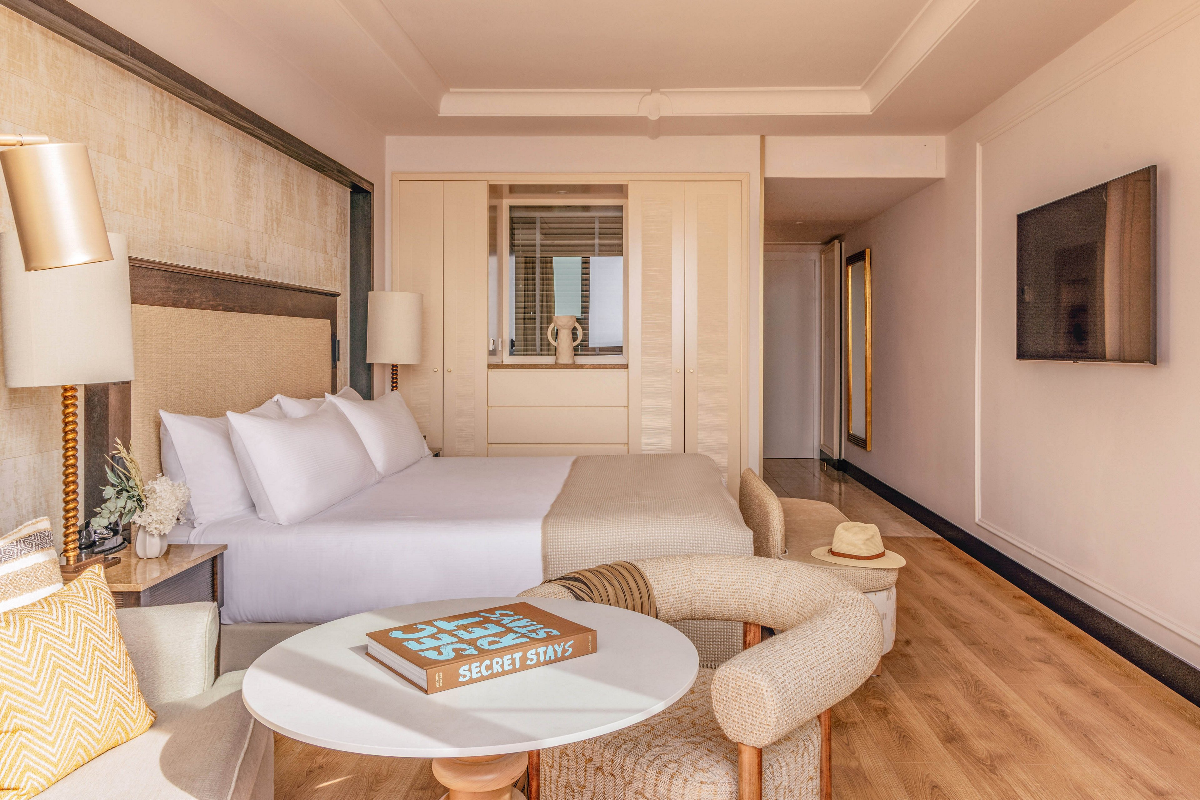 A spacious hotel room features a large neatly made bed with white linens and a padded headboard. In the foreground, a round table and upholstered chairs create a small lounge area, with a book titled "SECRET STAYS" placed on the tabletop. Warm wood flooring, soft neutral tones, and natural light from a window give the space an inviting atmosphere. A flat-screen television is mounted on the wall, and a straw hat rests casually on one of the chairs, suggesting a relaxed vacation setting.