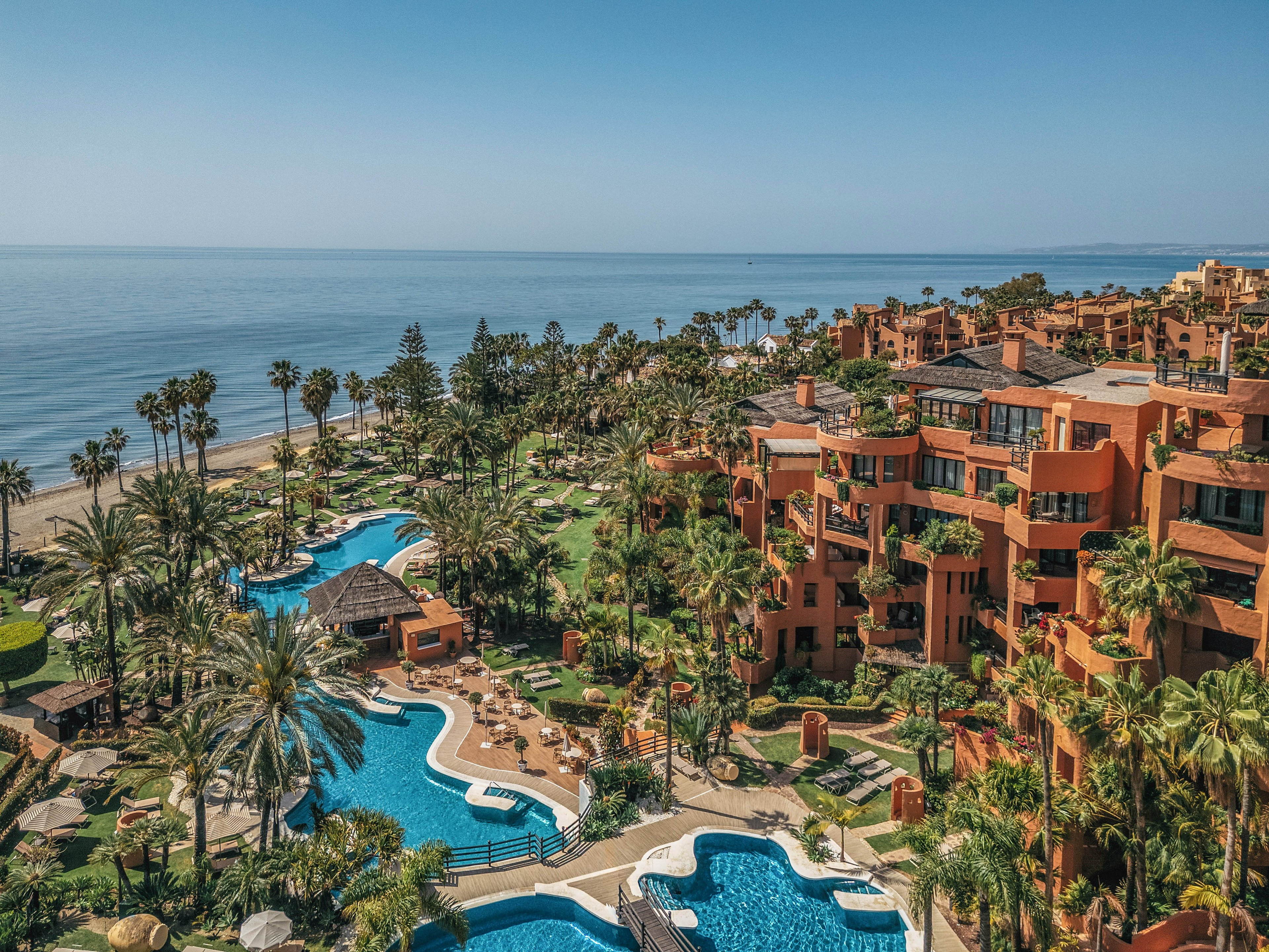A large seaside resort complex features multiple curving swimming pools surrounded by palm trees and lush greenery. Terracotta-colored apartment or hotel buildings rise along the right side, overlooking the pools and gardens. The scene opens onto a sandy beach and calm ocean under a clear blue sky. Sun loungers, pathways, and landscaped areas create a relaxed vacation atmosphere in a warm coastal destination.
