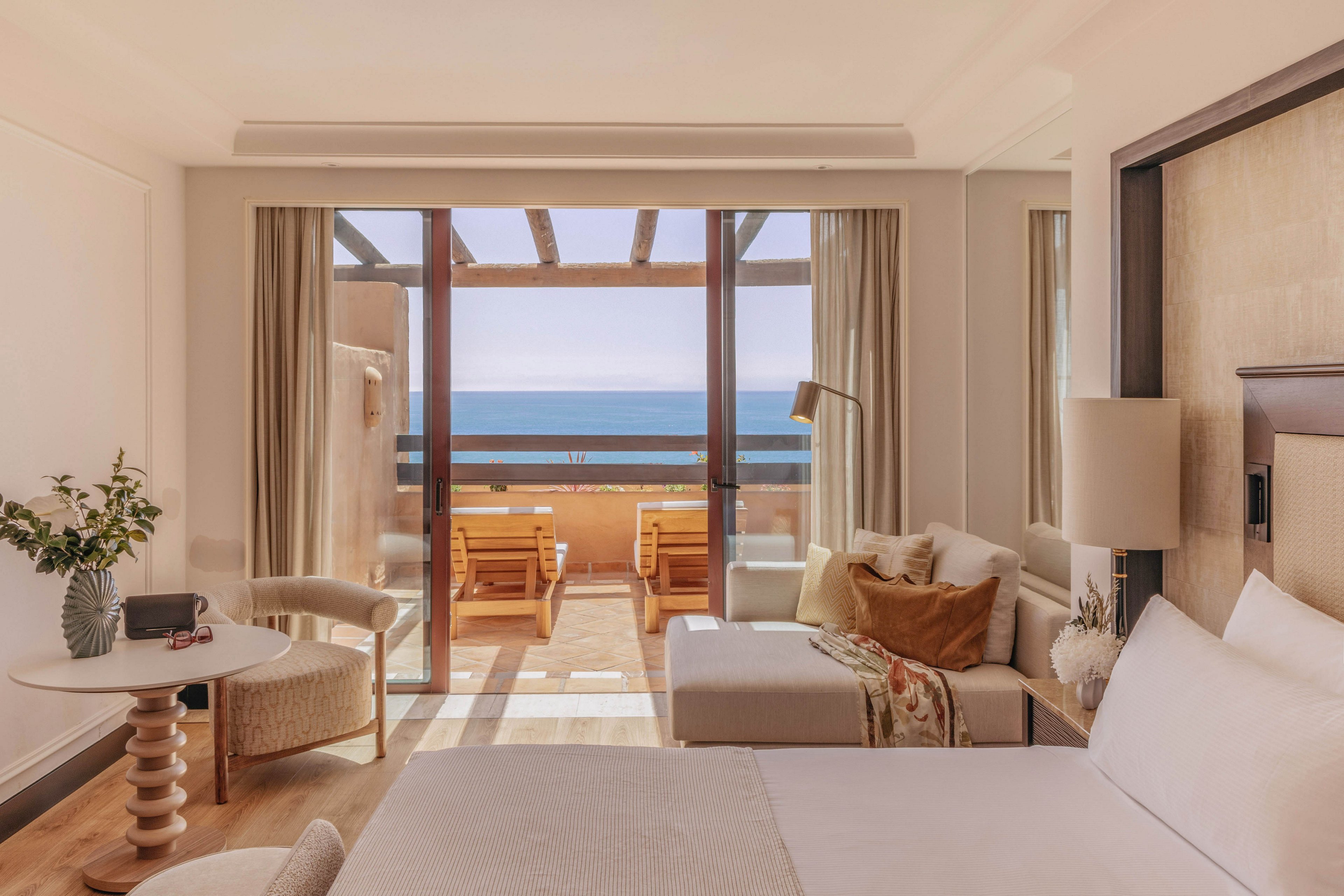 A bright, modern hotel room opens onto a balcony overlooking the ocean. The interior features a neatly made bed, a light-colored sofa, and a small round table with a vase of flowers. Large sliding glass doors let in abundant natural light and frame the sea view. Wooden balcony chairs and soft neutral tones create a calm, relaxing atmosphere ideal for a beachside getaway.