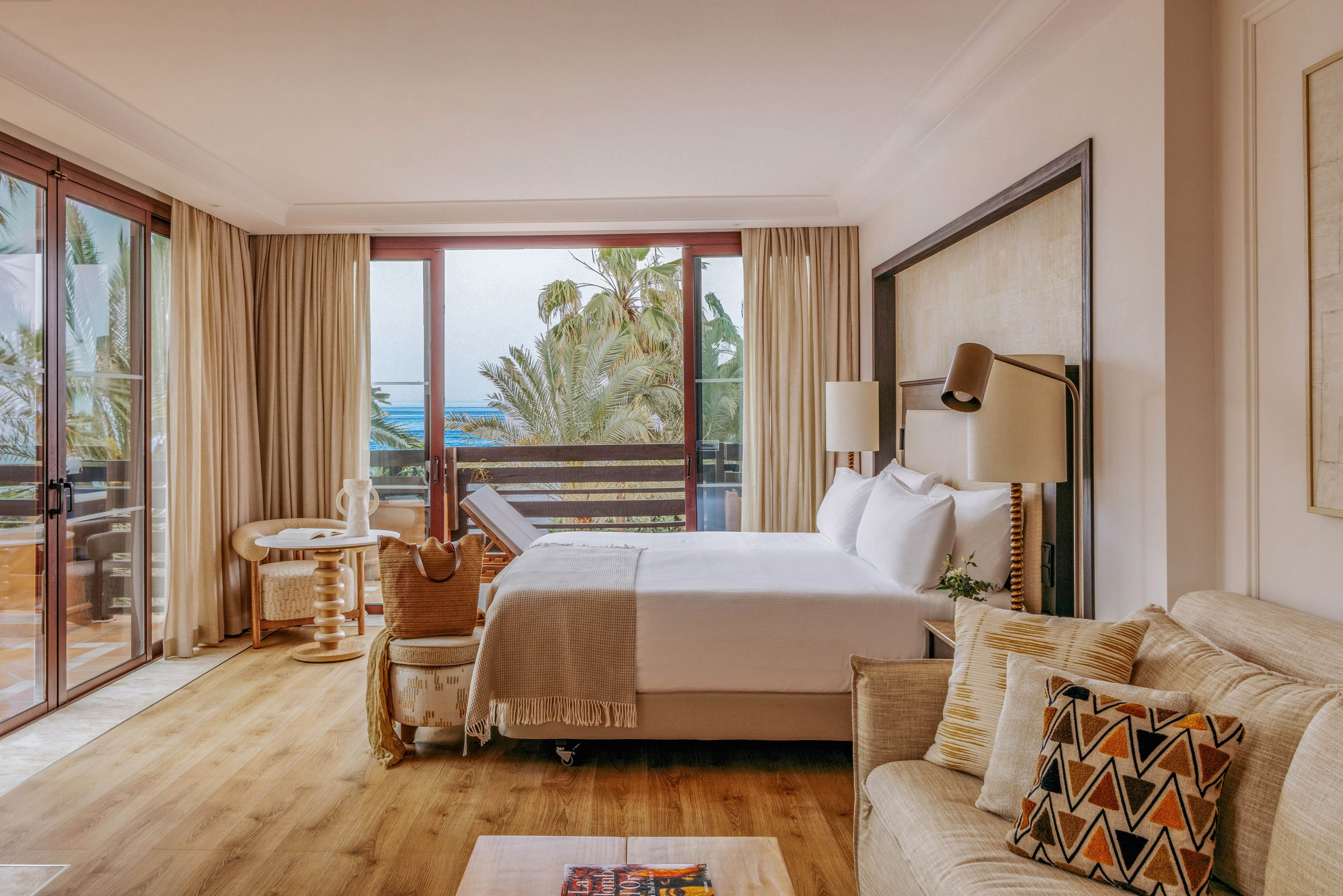 A spacious hotel bedroom features a neatly made double bed with neutral linens and soft pillows. Large floor-to-ceiling glass doors open to a balcony with views of palm trees and the sea. A small round table with chairs, a cozy sofa, and patterned cushions create a comfortable seating area. Warm wood flooring, beige curtains, and soft lighting give the room a calm, inviting atmosphere.