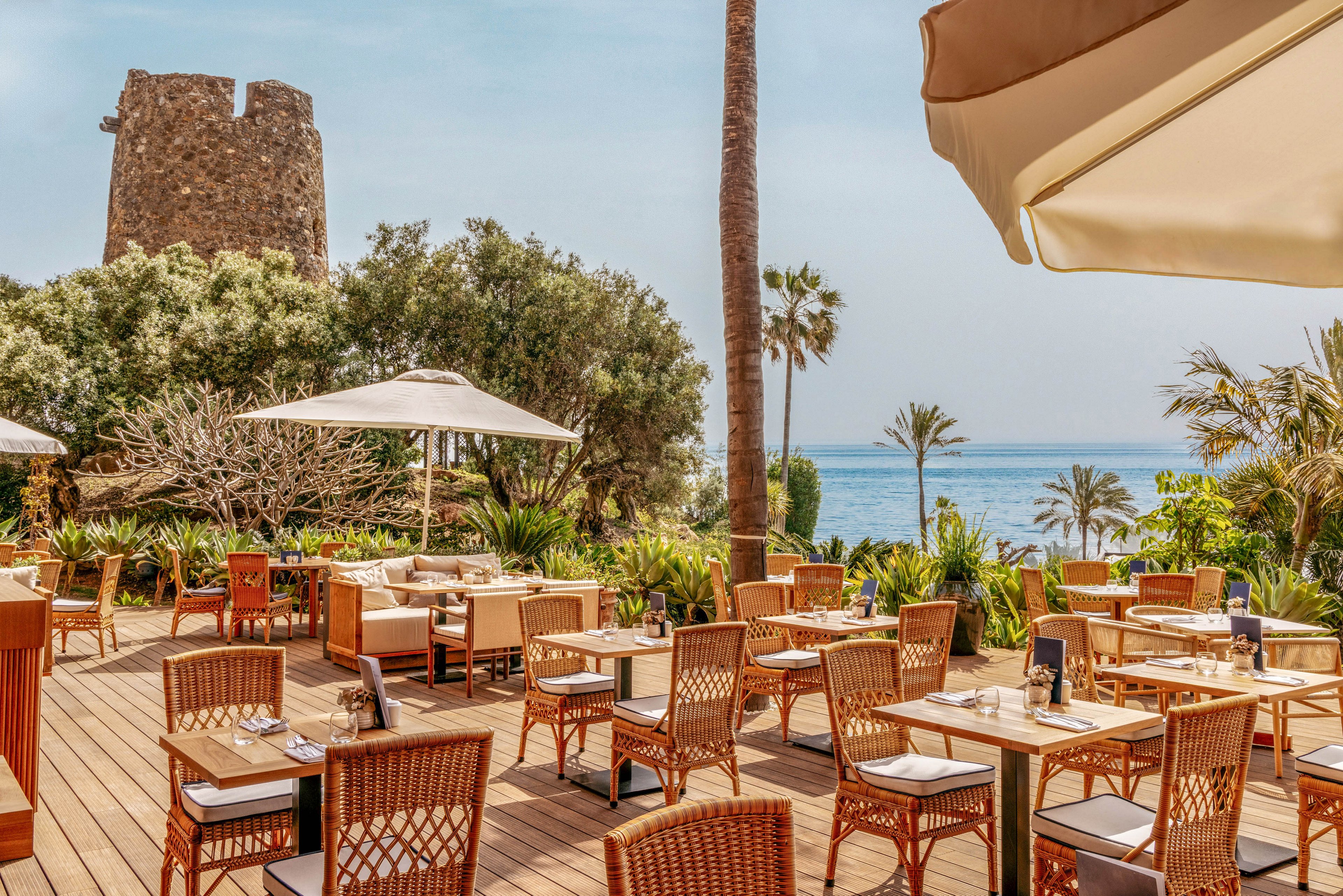 An open-air restaurant terrace features neatly arranged wooden tables and wicker chairs under large white umbrellas. The setting overlooks lush greenery and a calm blue sea in the background. A historic stone tower rises behind the dining area, adding a distinctive landmark to the coastal scene. Warm sunlight and natural materials create a relaxed, inviting atmosphere for outdoor dining.