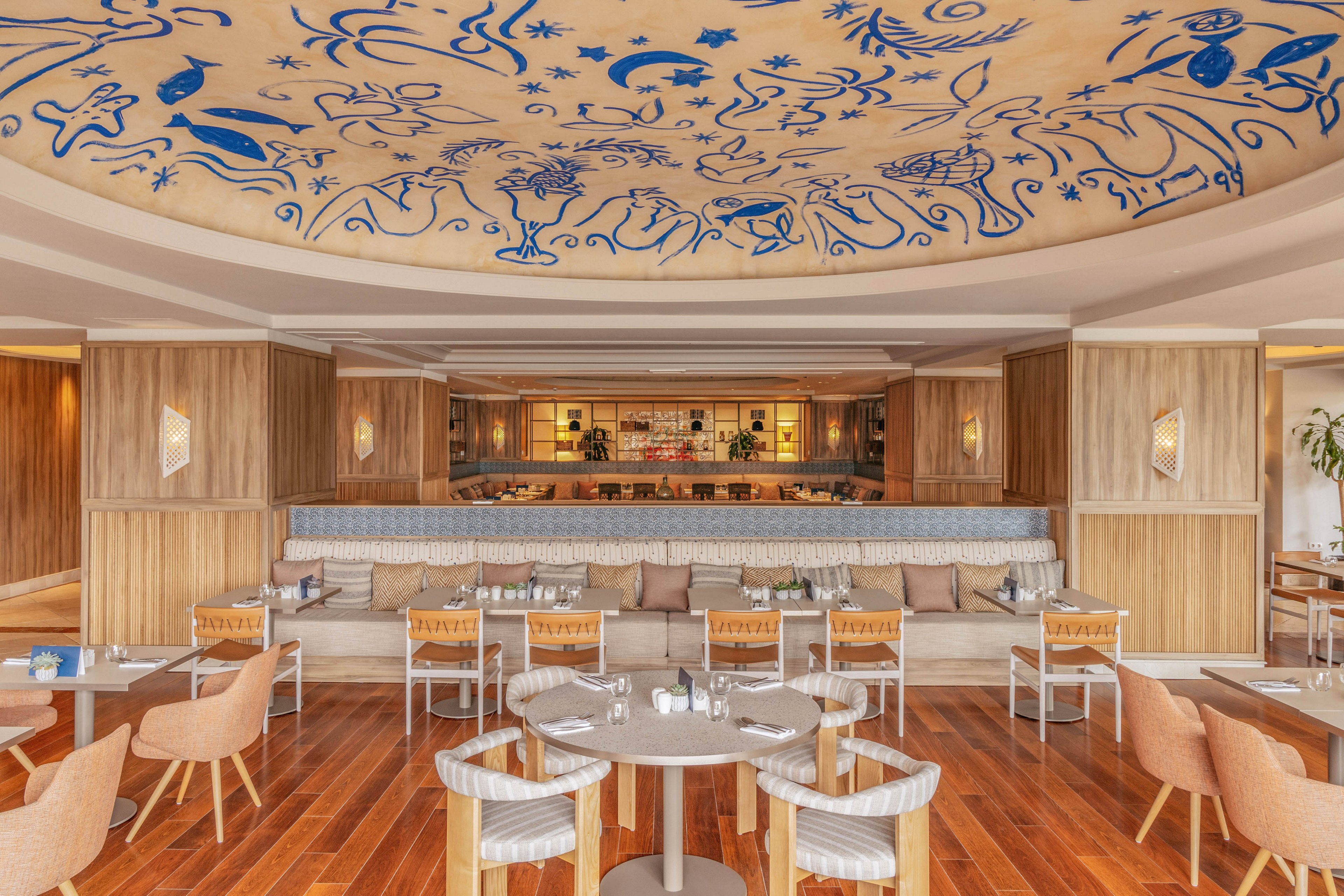Contemporary lounge bar interior features light wood finishes and a central bar at the back. The space includes cushioned bench seating, round dining tables, and upholstered chairs arranged on polished wooden flooring. A large decorative ceiling with blue abstract illustrations and symbols creates a striking focal point. Warm ambient lighting and neutral tones give the room a relaxed, upscale atmosphere.