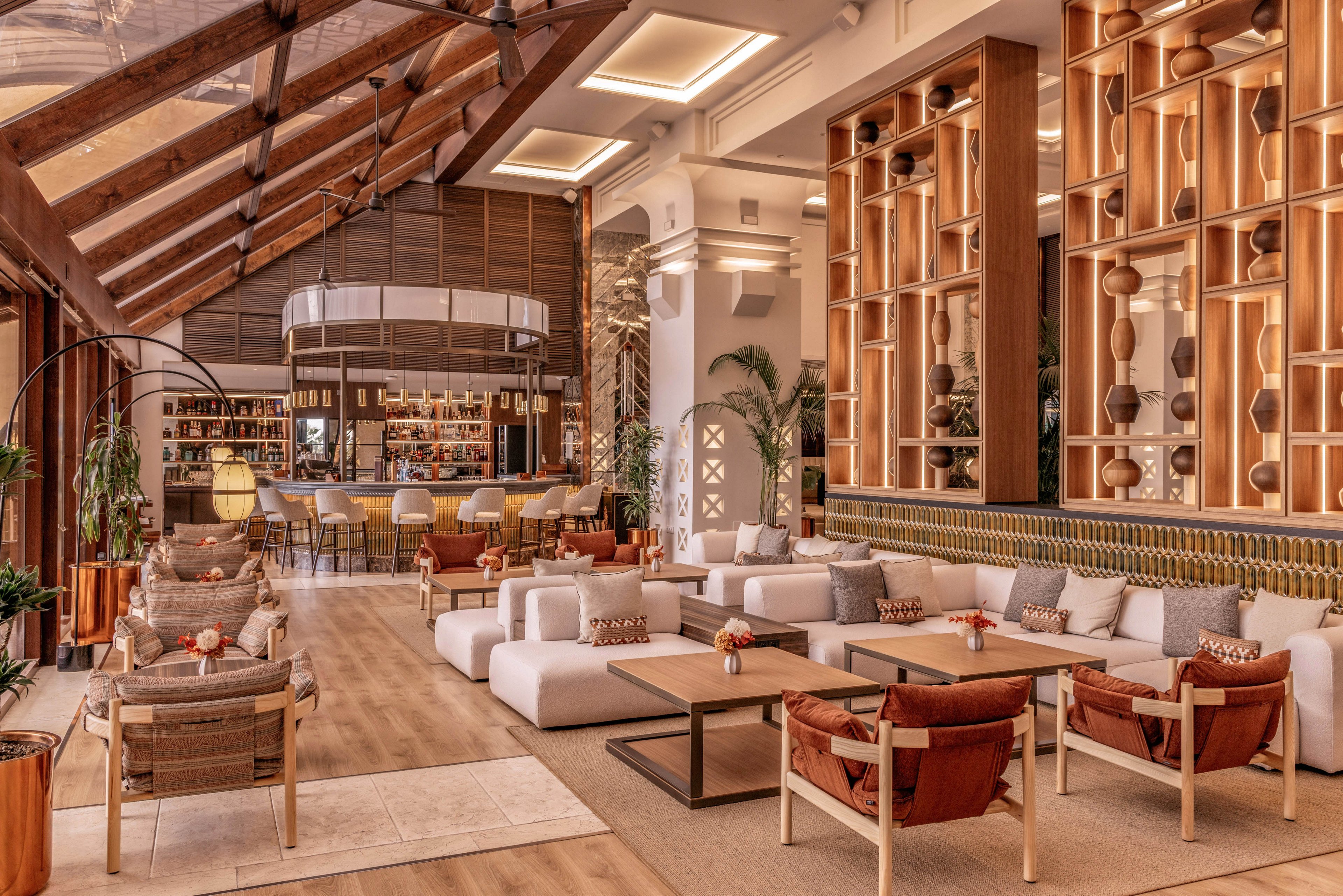 Spacious hotel lounge features modern seating areas with plush white sofas and wooden chairs. A bar is visible in the background, framed by warm lighting and shelves of bottles. Large floor-to-ceiling windows and wooden accents create an airy, sunlit atmosphere. Decorative vases, cushions, and plants add texture and a welcoming, upscale feel to the interior.