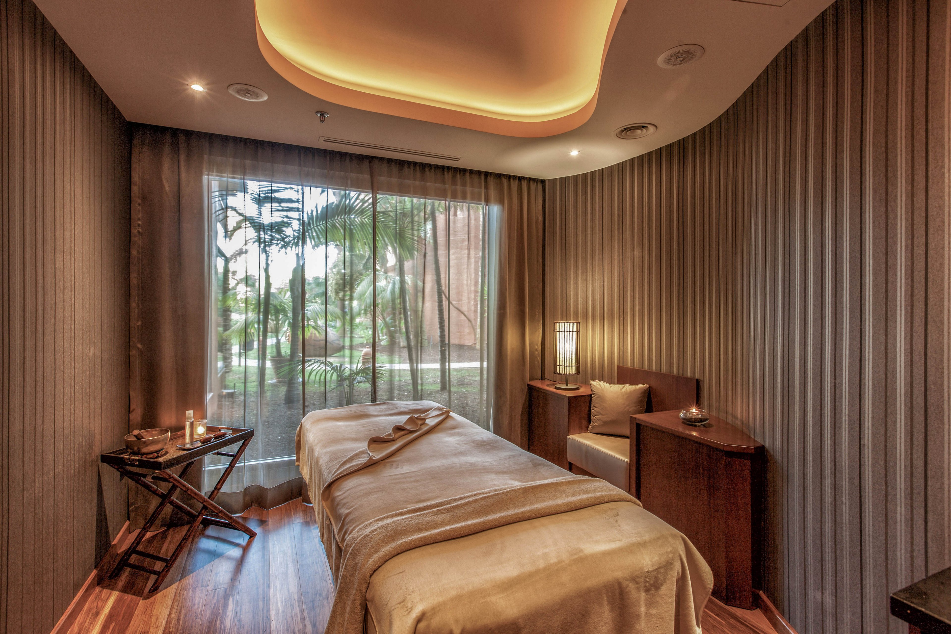 A neatly made massage table stands in the center of a cozy spa treatment room. Soft, warm lighting from the ceiling and a table lamp creates a relaxing atmosphere. Sheer curtains reveal a view of lush green palm trees outside the window. Candles and neatly arranged towels on a side table enhance the tranquil, wellness-focused setting.