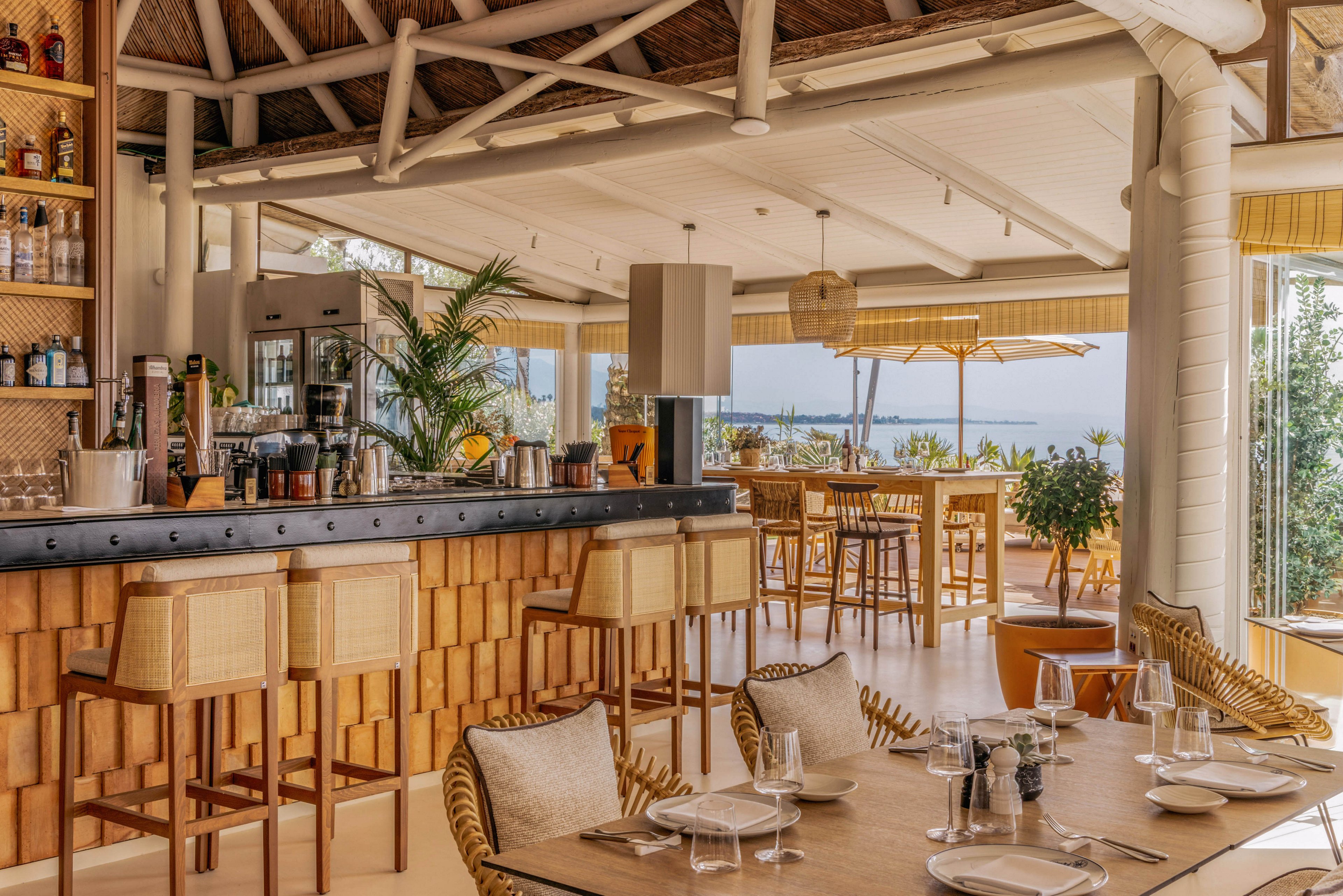 An inviting open-air bar and dining area overlooks a bright coastal landscape. Wooden bar stools and woven chairs surround neatly set tables with glassware and plates. Natural materials, warm beige tones, and soft lighting create a relaxed, beachside atmosphere. Large windows and an outdoor terrace provide views of the water and distant shoreline.