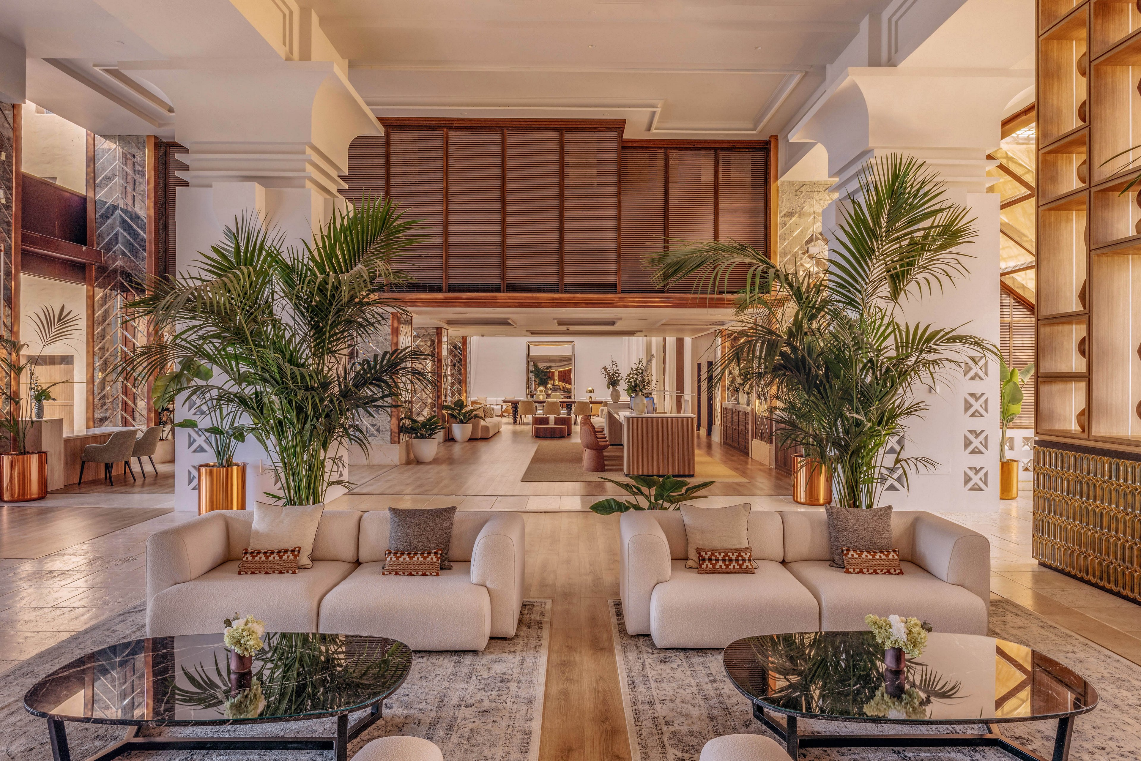 A large, elegant hotel lobby features modern cream-colored sofas arranged around dark glass coffee tables. Tall green palm plants in copper-toned planters frame the central walkway and add a tropical feel. Warm wood accents, soft lighting, and neutral rugs create a welcoming, upscale atmosphere. The open layout leads the eye toward a reception or lounge area in the background.