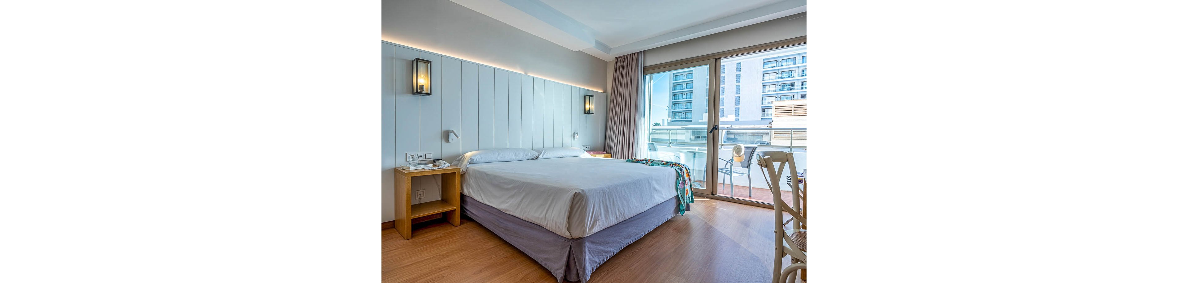 Bright contemporary hotel room featuring a neatly made double bed with white linens and a light gray bed skirt. The space includes wooden bedside tables, wall-mounted lamps, and soft neutral wall paneling. Large sliding glass doors open to a balcony overlooking nearby apartment buildings, allowing natural light to fill the room. A colorful blanket adds a touch of accent color to the otherwise minimalist, clean interior.