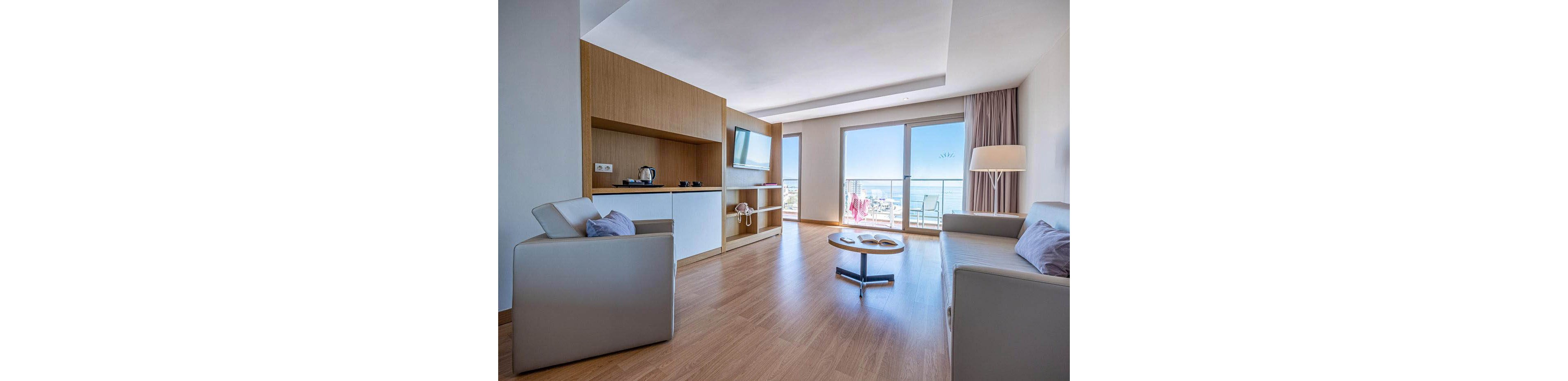 Bright contemporary hotel suite features a seating area with armchairs and a sofa around a low round coffee table. A built-in wooden kitchenette with a kettle and cups sits opposite a wall-mounted television. Large sliding glass doors open to a balcony overlooking the ocean and outdoor seating. Soft neutral tones, clean lines, and natural light create a calm, minimalist atmosphere.