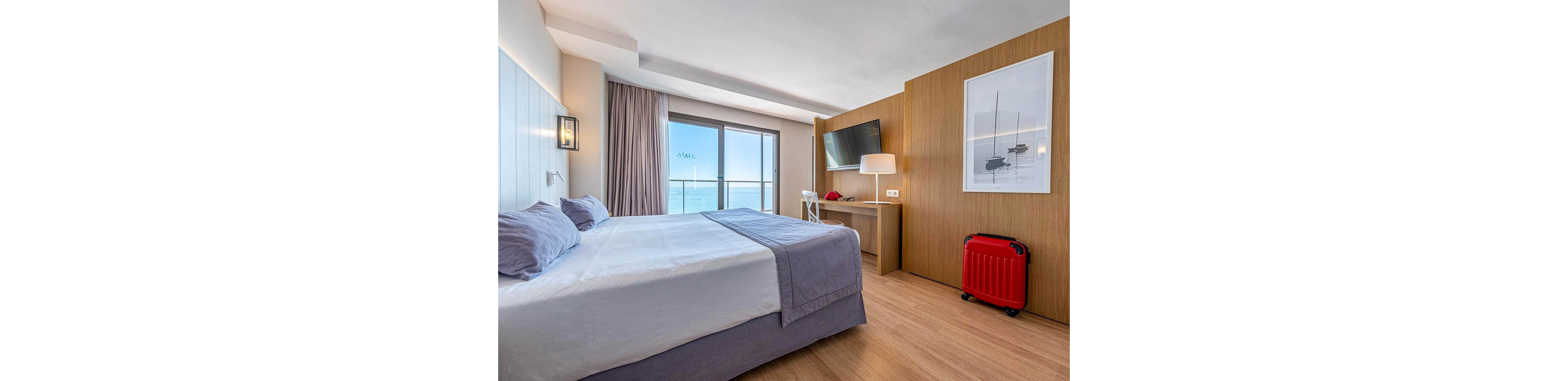 A contemporary hotel room features a neatly made double bed with light bedding and soft pillows. Large glass doors open to a balcony overlooking the ocean, allowing natural light to fill the space. A wall-mounted television, desk with chair, and floor lamp create a comfortable area for work or relaxation. A bright red suitcase stands near the wooden wall, adding a pop of color to the calm, neutral interior.