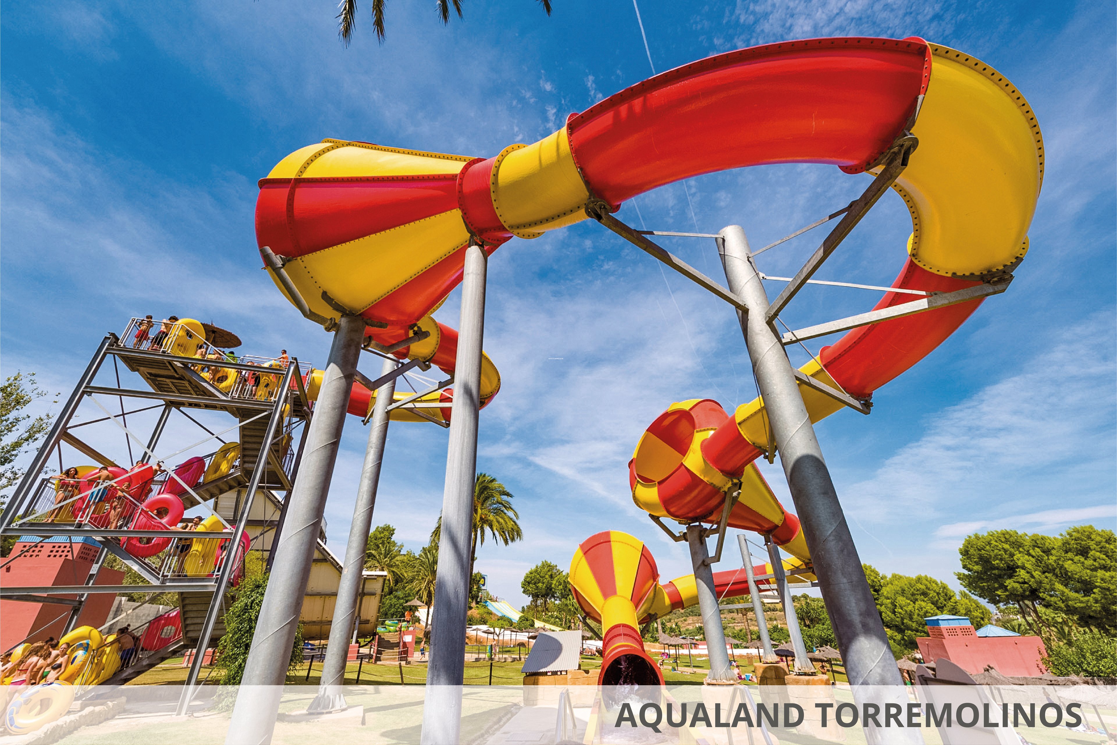 A vibrant outdoor water park featuring large red and yellow water slides under a clear blue sky. The setting includes lush greenery and palm trees, creating a tropical atmosphere. The text 'Aqualand Torremolinos' is visible in the image, identifying the location.