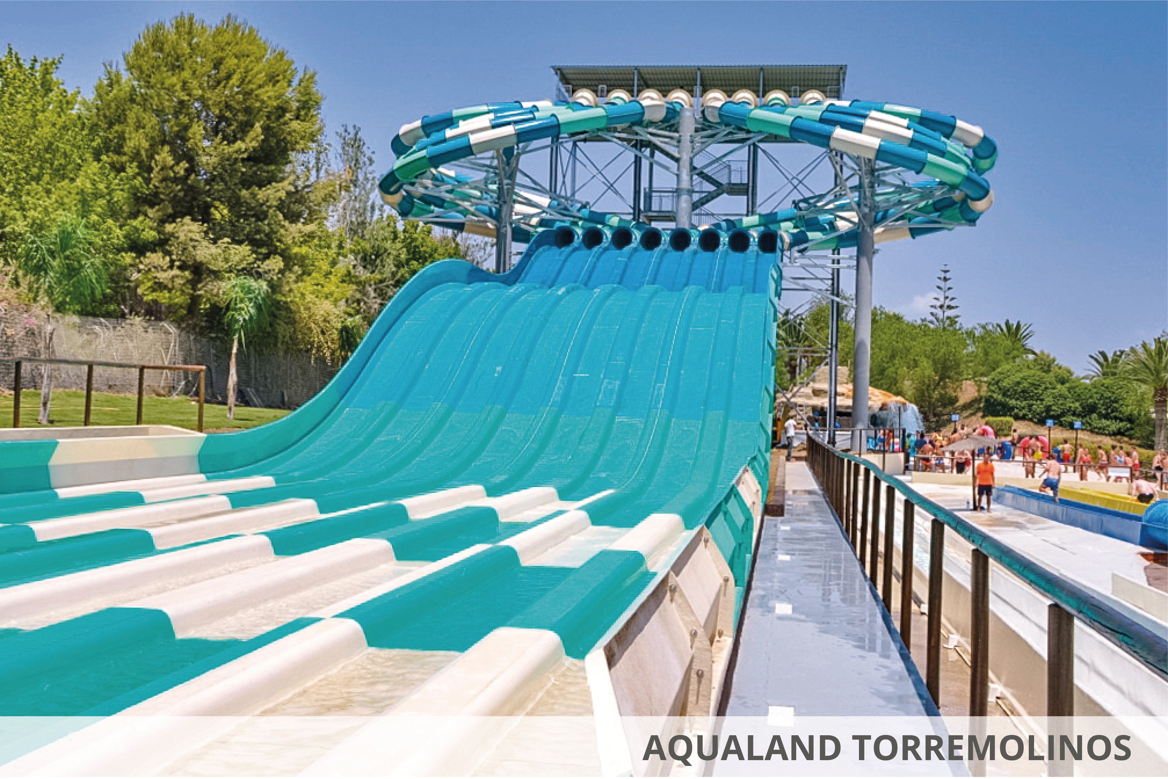 A vibrant multi-lane water slide at Aqualand Torremolinos, set in a sunny outdoor environment surrounded by greenery. The slide features alternating teal and white lanes, leading to a pool area. Visitors can be seen enjoying the park in the background, adding to the lively atmosphere.