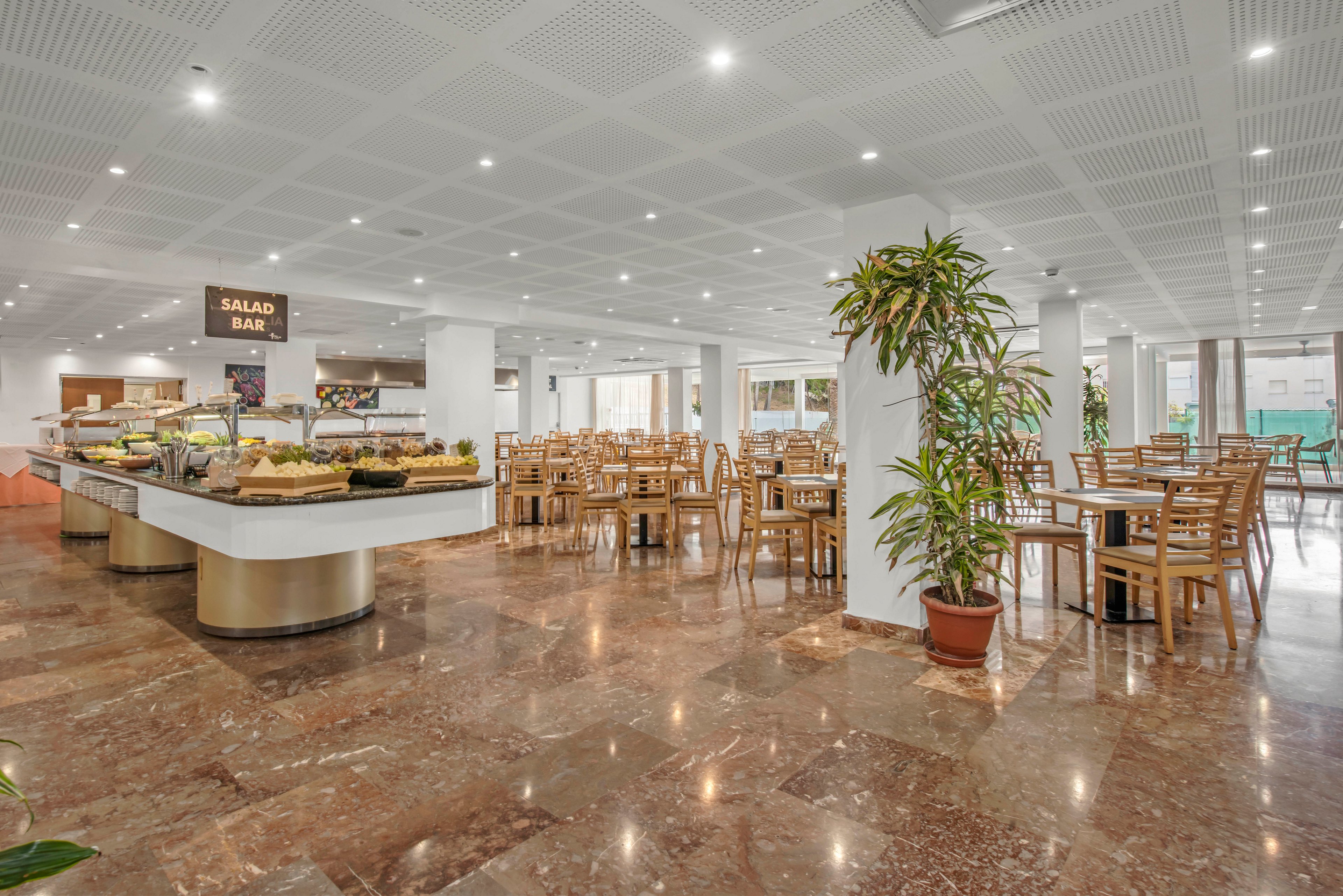 Spacious self-service restaurant interior featuring a central salad bar filled with fresh food trays. Wooden tables and chairs are neatly arranged across the polished stone floor, creating an organized dining area. Large windows and bright ceiling lights provide ample natural and artificial light, giving the space an airy, modern feel. A sign reading "SALAD BAR" clearly marks the buffet section in the middle of the room.