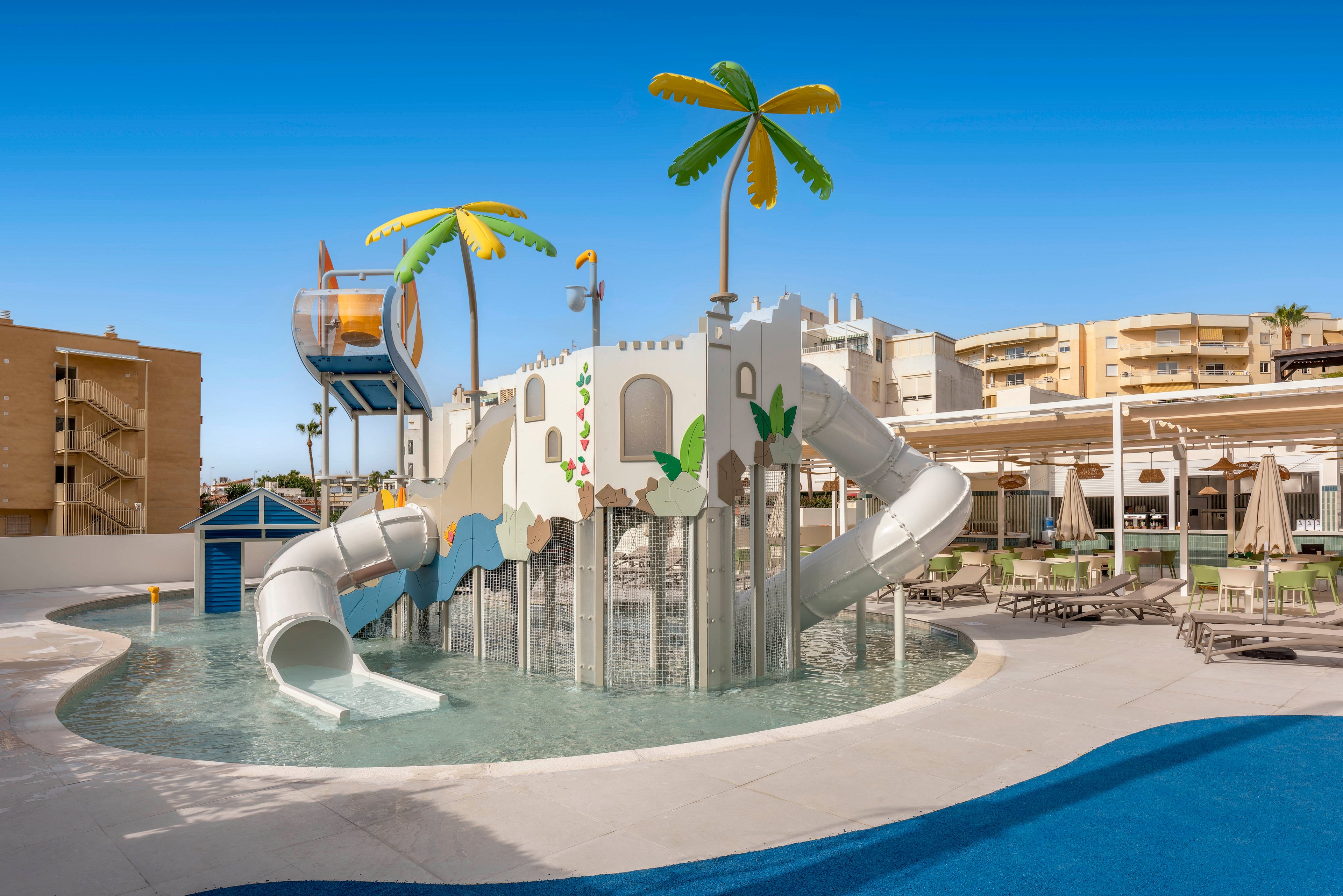 A bright resort water playground features multiple white slides and a castle-like structure set in a shallow pool. Decorative palm trees and playful shapes add a fun, tropical theme to the area. Sun loungers and umbrellas surround the pool, suggesting a family-friendly vacation setting. Apartment-style buildings and clear blue sky frame the scene in the background.