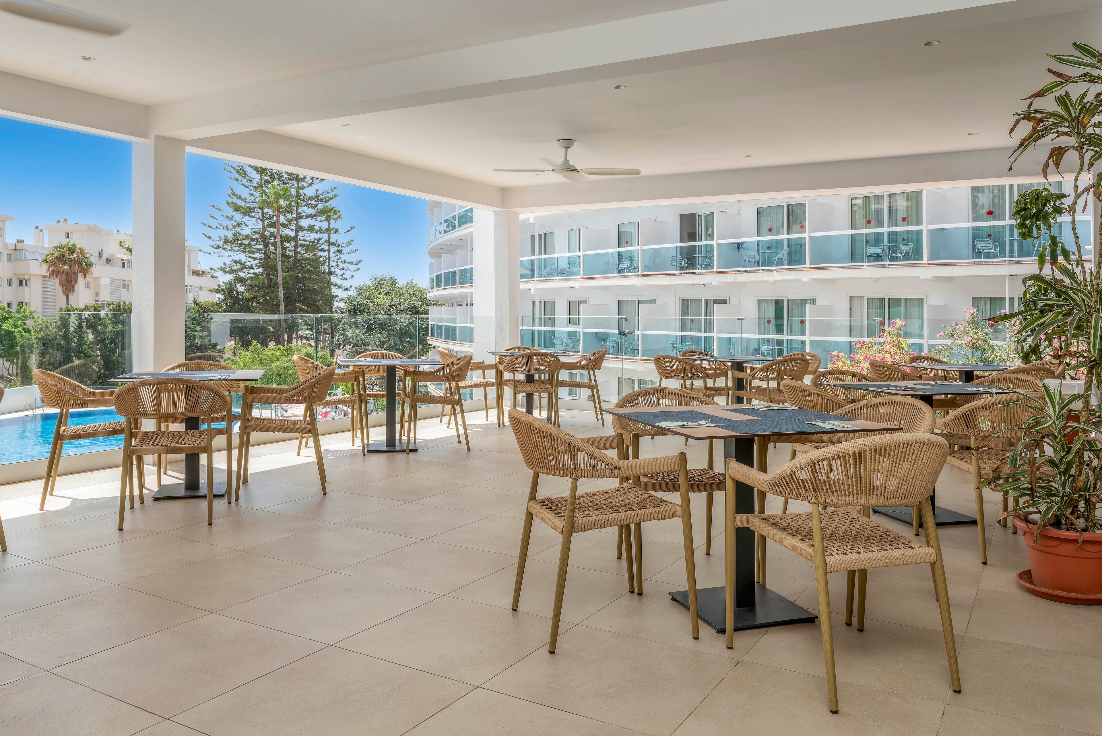 Open-air hotel terrace features several tables with wicker chairs arranged neatly on a tiled floor. The space overlooks a swimming pool and a modern multi-story building with glass balconies. Large plants in pots add greenery to the bright, minimalist setting. Clear blue sky and surrounding trees create a relaxed, vacation atmosphere.