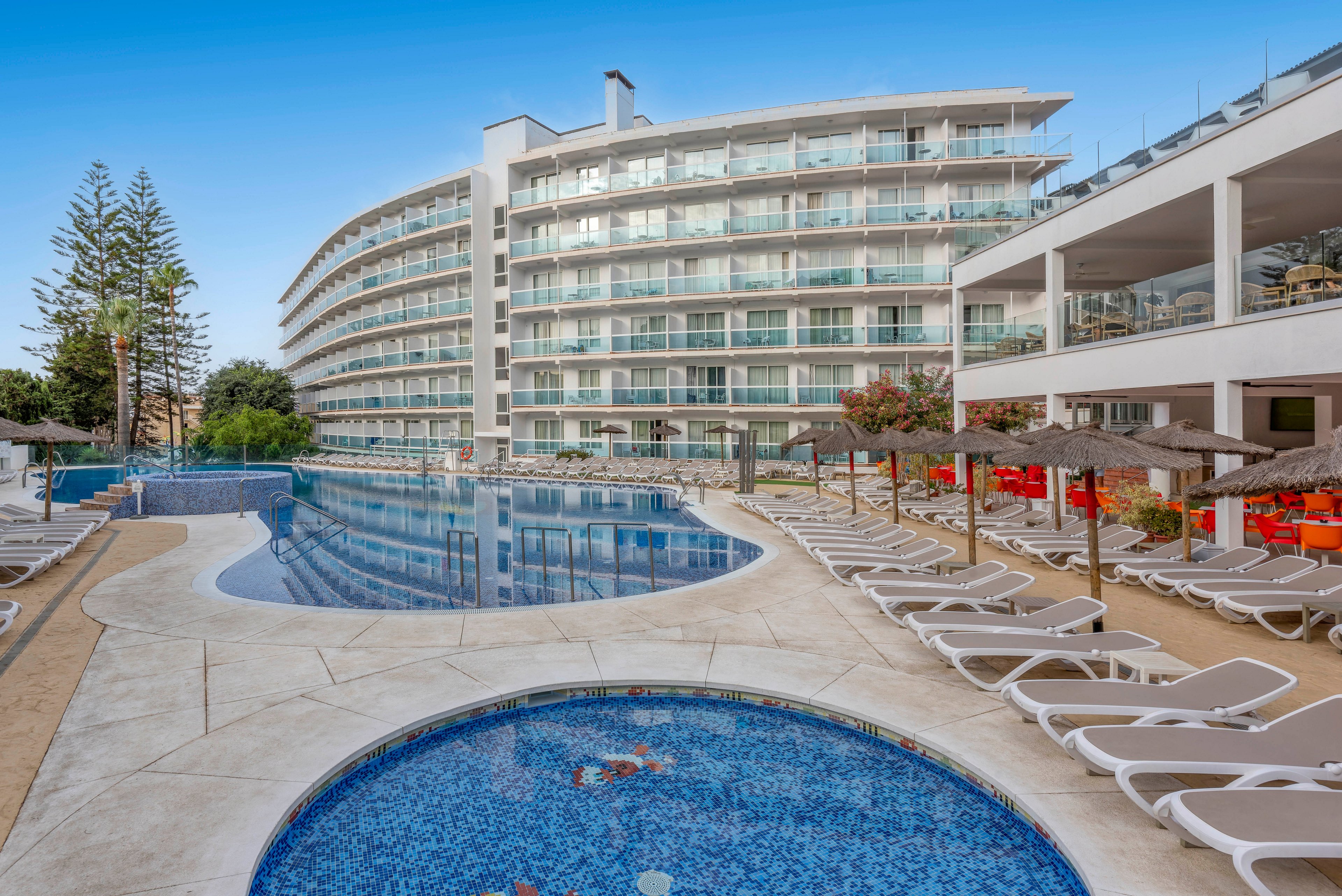 A contemporary multi-story resort hotel overlooks a large curved outdoor swimming pool and a smaller round children's pool in the foreground. Numerous white sun loungers line the poolside terraces, creating a relaxed vacation atmosphere. The building features glass balconies on each floor, with trees and greenery visible around the property. Clear blue sky and calm water reflections emphasize a bright, sunny holiday setting.