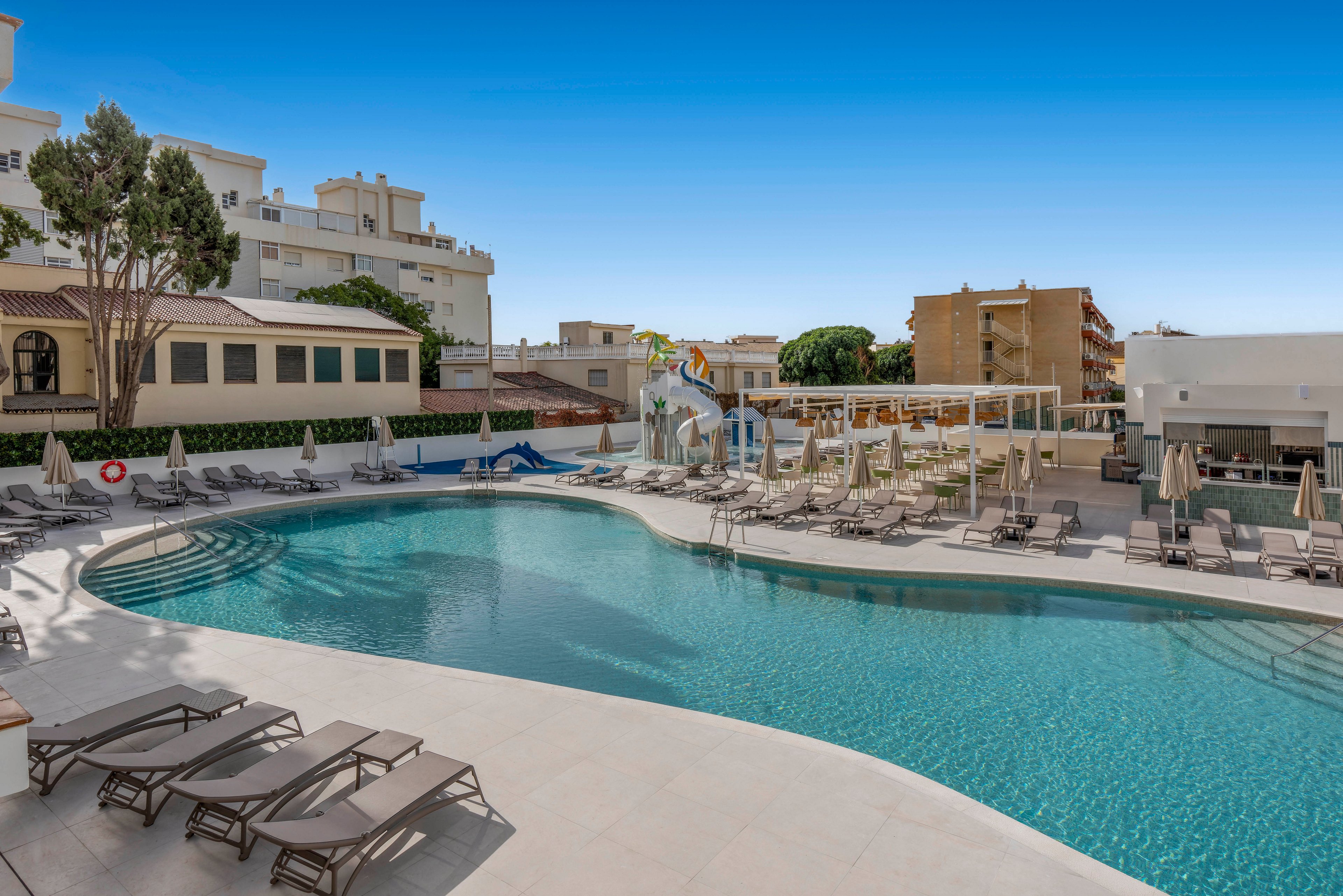 A large outdoor swimming pool is surrounded by numerous sun loungers and umbrellas at a modern resort. Apartment-style buildings and trees frame the pool area under a clear blue sky. A small water slide and play structure are visible near the far side of the pool, suggesting family-friendly amenities. The scene conveys a relaxed vacation atmosphere with clean, contemporary architecture and inviting turquoise water.