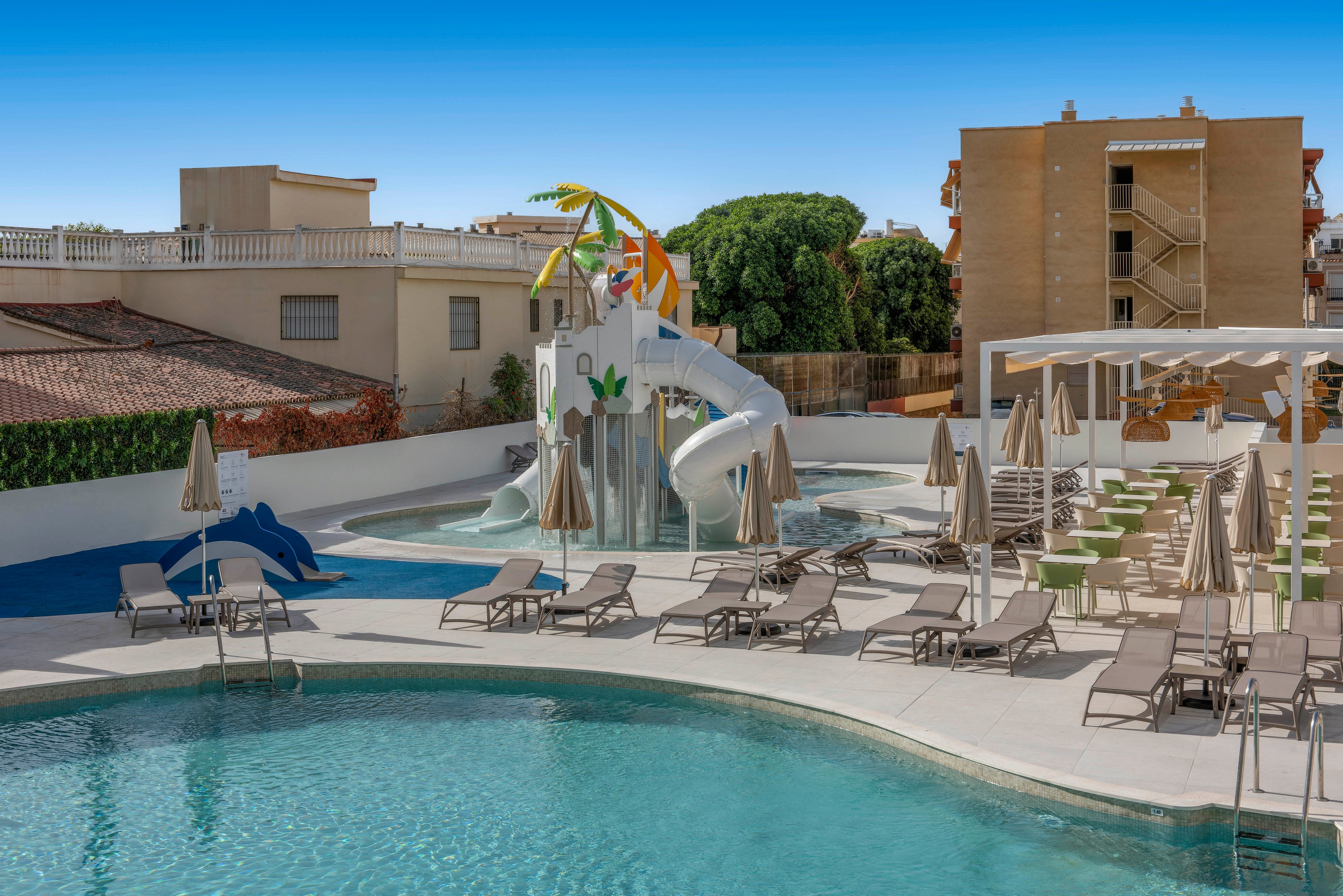 Outdoor resort swimming pool area featuring a playful waterslide structure with colorful decorations. Numerous sun loungers and beige umbrellas surround the pool, creating a relaxed vacation setting. Nearby buildings and trees frame the scene under a clear blue sky. The space appears calm and empty, emphasizing leisure and family-friendly recreation.