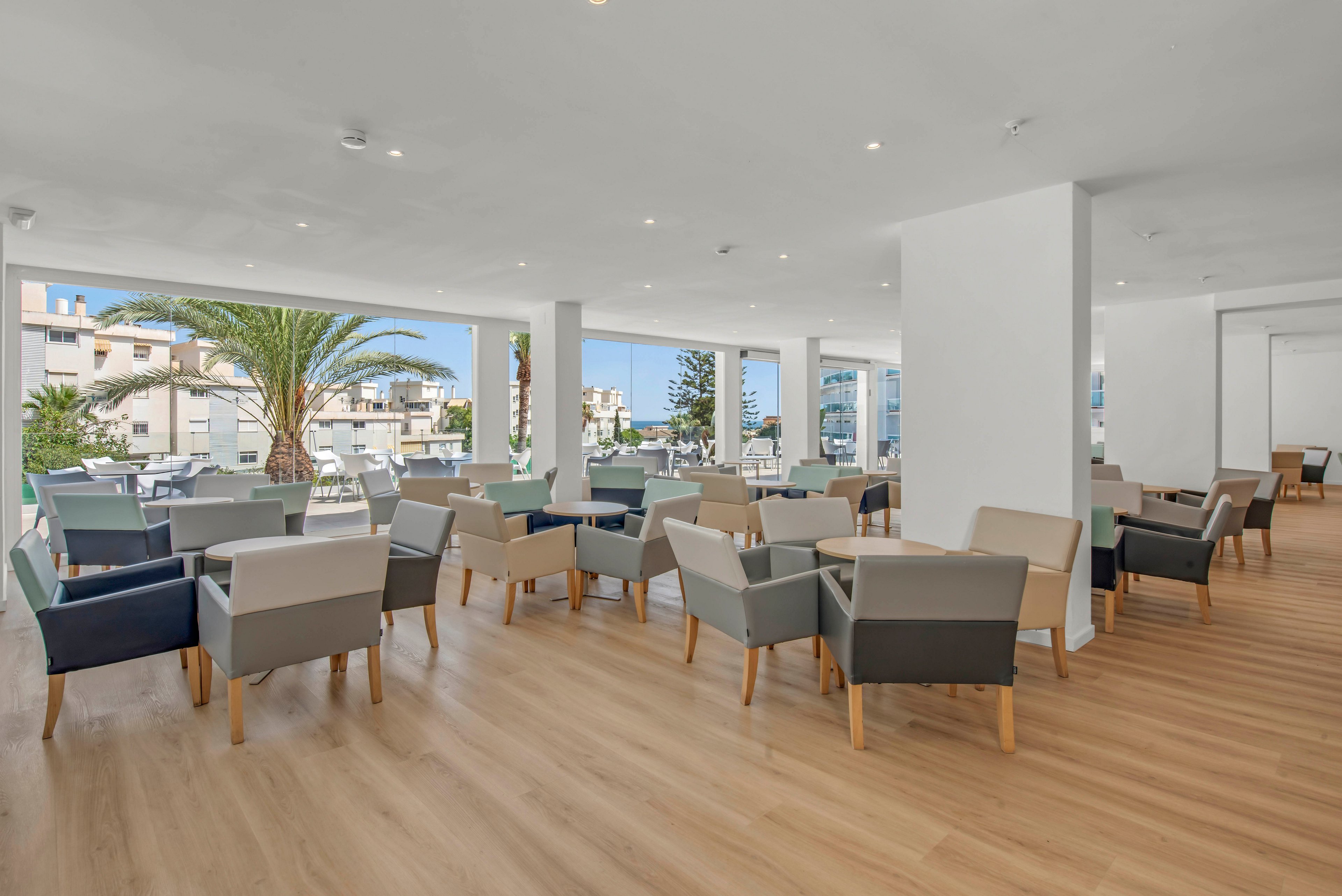 Spacious indoor lounge area featuring rows of modern armchairs and small tables on a light wood floor. Large open windows reveal a sunny coastal town with palm trees and white buildings outside. The space is filled with natural daylight, creating an airy and relaxed atmosphere. Neutral-toned furniture and clean architectural lines emphasize a contemporary, minimalist style.