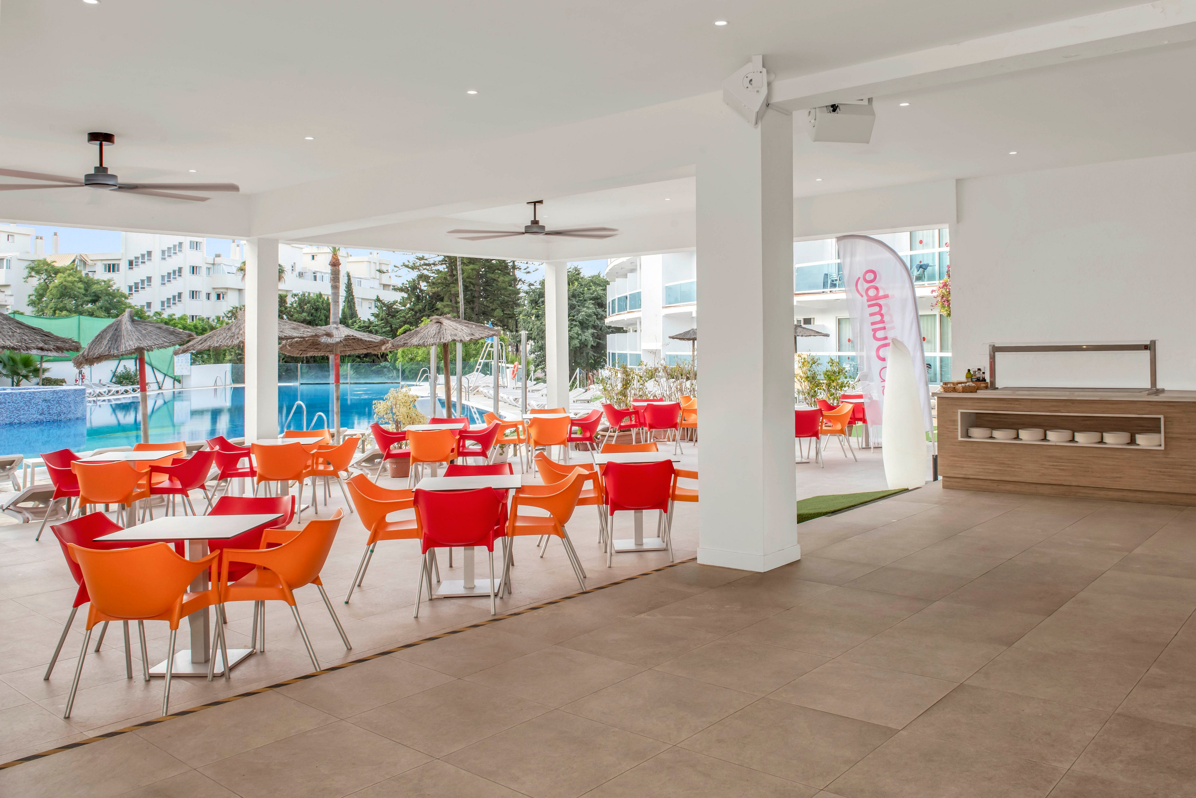 An open-air cafe area features bright orange and red chairs around white tables under a covered terrace. The space overlooks a blue swimming pool with thatched umbrellas and surrounding modern white buildings. A tall white flag with red text stands near the seating area, adding a touch of branding to the scene. The overall atmosphere is clean, relaxed, and inviting for guests enjoying a poolside break.