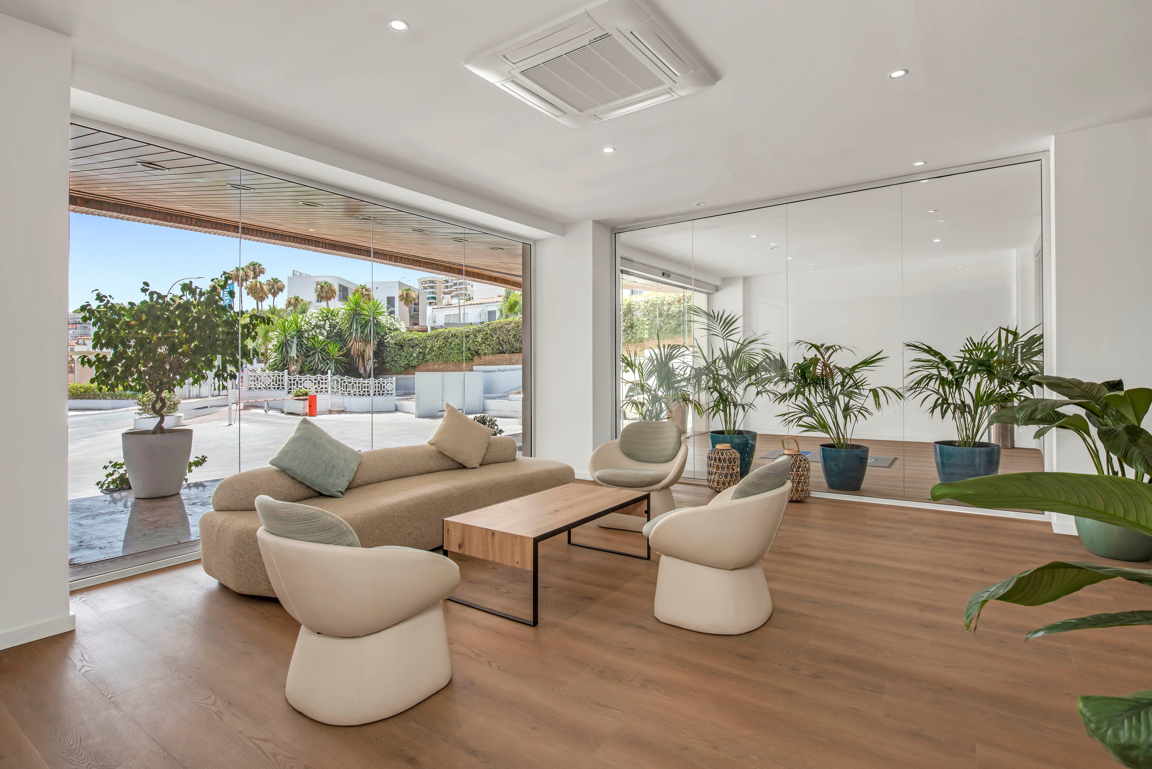 Contemporary lounge area featuring a beige sofa and three sculptural armchairs around a wooden coffee table. Floor-to-ceiling glass windows open to an outdoor view with plants and a sunny street. Potted greenery lines the interior wall, adding a natural touch to the minimalist decor. Light wood flooring and white walls create an airy, spacious atmosphere suitable for reception or waiting area use.