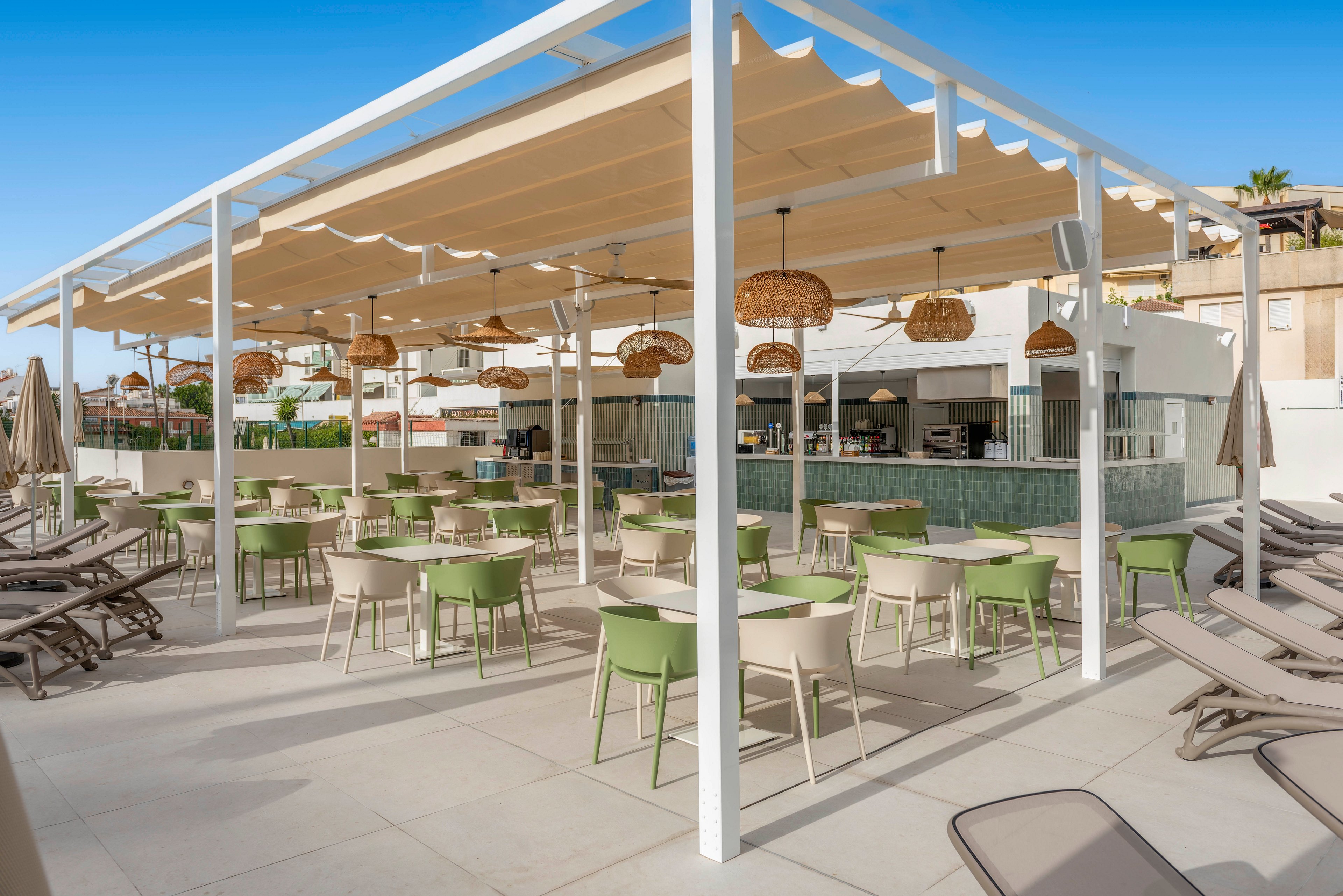 Open-air terrace bar area with green chairs and white tables is arranged beneath a large pergola structure. Wicker pendant lights hang from the ceiling, adding a warm decorative touch to the modern setting. Sun loungers line the surrounding space, suggesting a poolside or resort environment under a clear blue sky. Buildings and umbrellas in the background complete the relaxed vacation atmosphere.