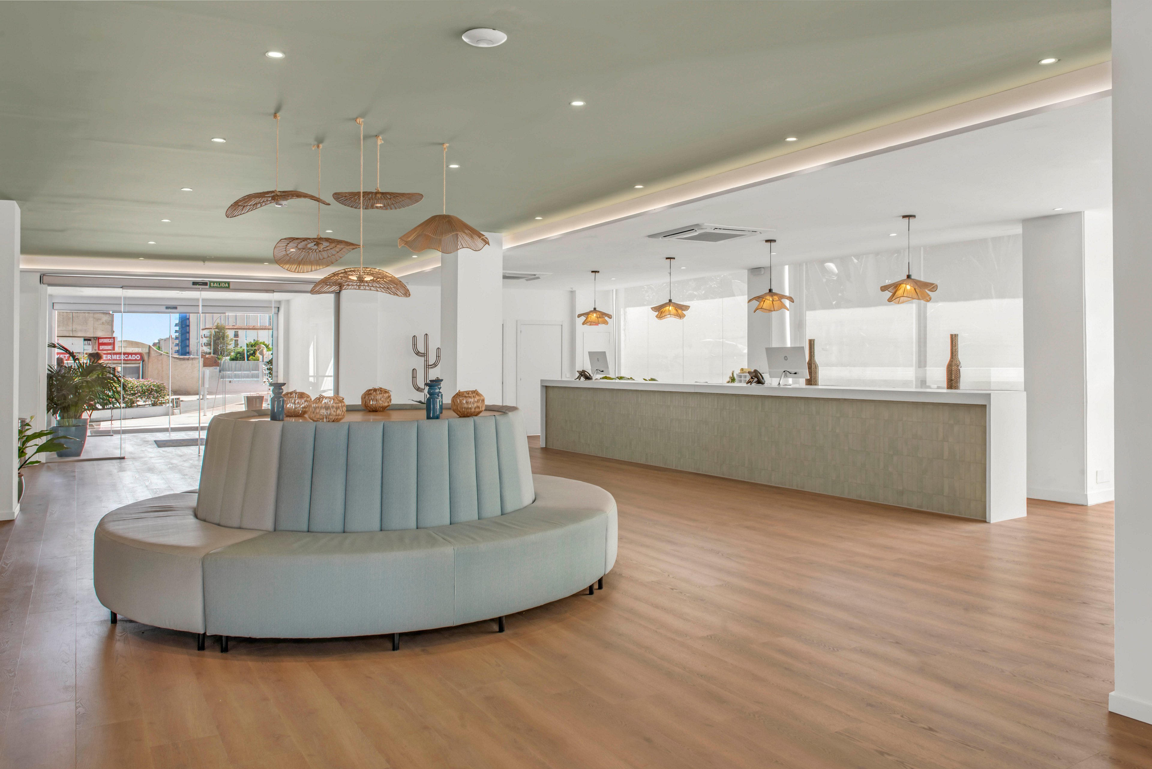 Spacious contemporary lobby interior featuring a large circular upholstered bench in soft pastel tones. A long minimalist reception desk with pendant lights runs along one side of the room. Wooden flooring and a light-colored ceiling create a warm, inviting atmosphere. Decorative plants and sculptural objects add visual interest near the entrance and seating area.