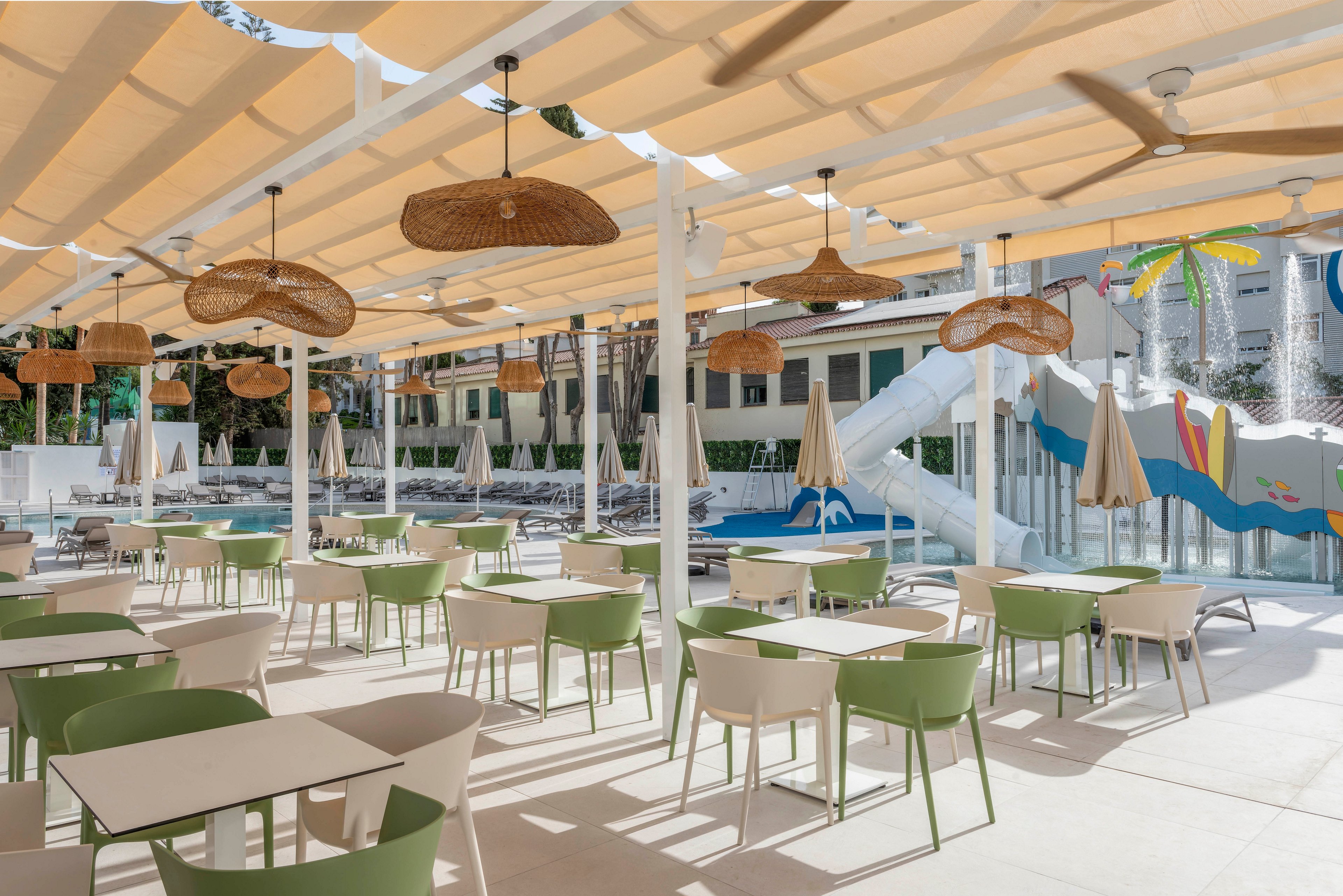 Bright outdoor seating area with modern tables and green chairs is set under a large pergola with fabric shades and woven pendant lights. The café overlooks a poolside zone featuring sun loungers, beige umbrellas, and a colorful water slide structure. White columns, ceiling fans, and clean architectural lines create a relaxed, contemporary resort atmosphere. The scene is empty of people, emphasizing the tranquil, sunny setting.