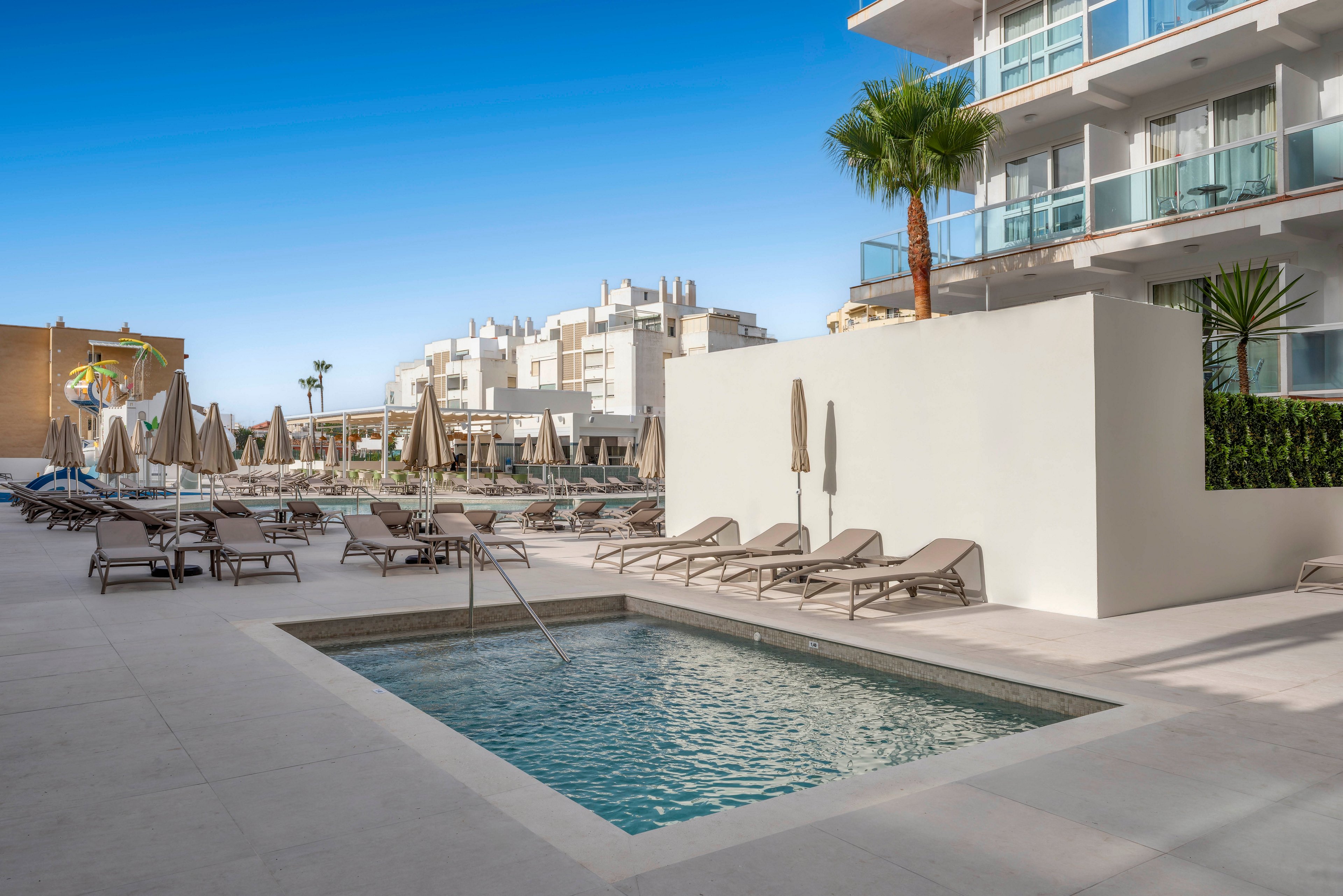 A small rectangular pool sits at the center of a modern hotel terrace surrounded by beige sun loungers and closed umbrellas. The setting appears to be an urban resort with multi-story balconies overlooking the pool area. Clean architectural lines, light stone flooring, and minimalistic design create a contemporary, relaxed atmosphere. Clear blue sky and palm trees add a warm, vacation feel to the scene.