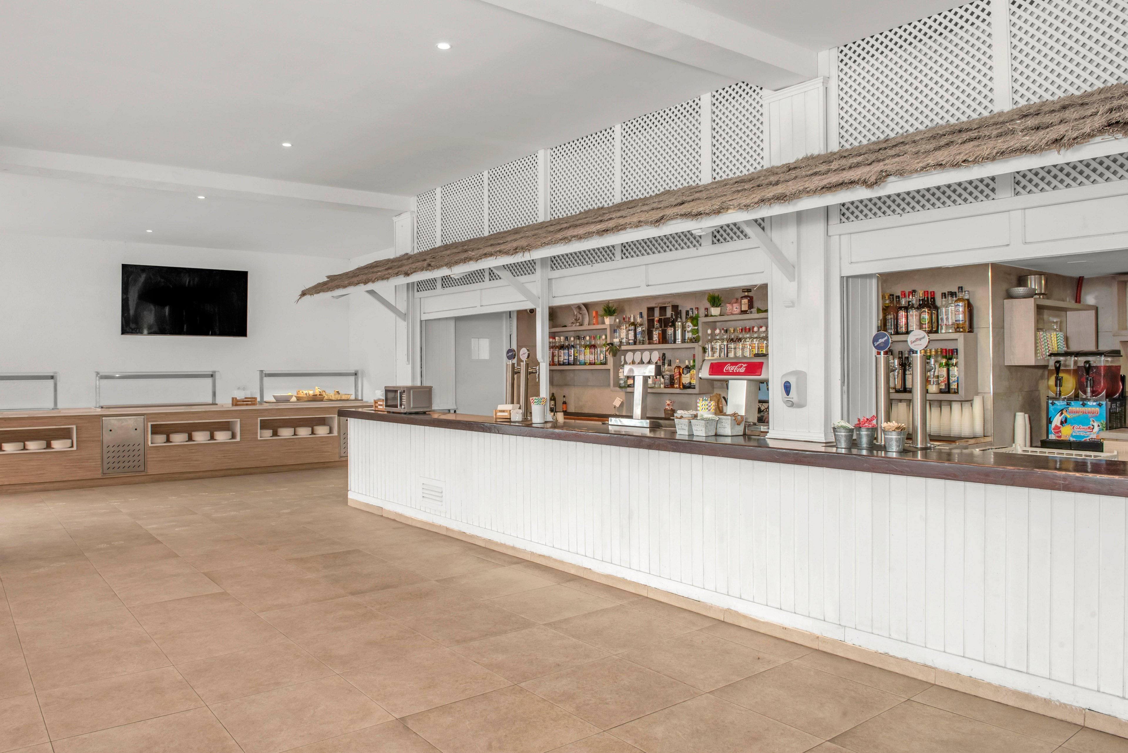 Spacious bar interior featuring a long white counter and tiled floor in a contemporary resort setting. Shelves behind the bar display various bottles and drink dispensers, with bar equipment neatly arranged. A large flat-screen television is mounted on the wall opposite the open floor area. Neutral tones and clean lines create a bright, minimalist atmosphere suitable for hospitality or leisure environments.