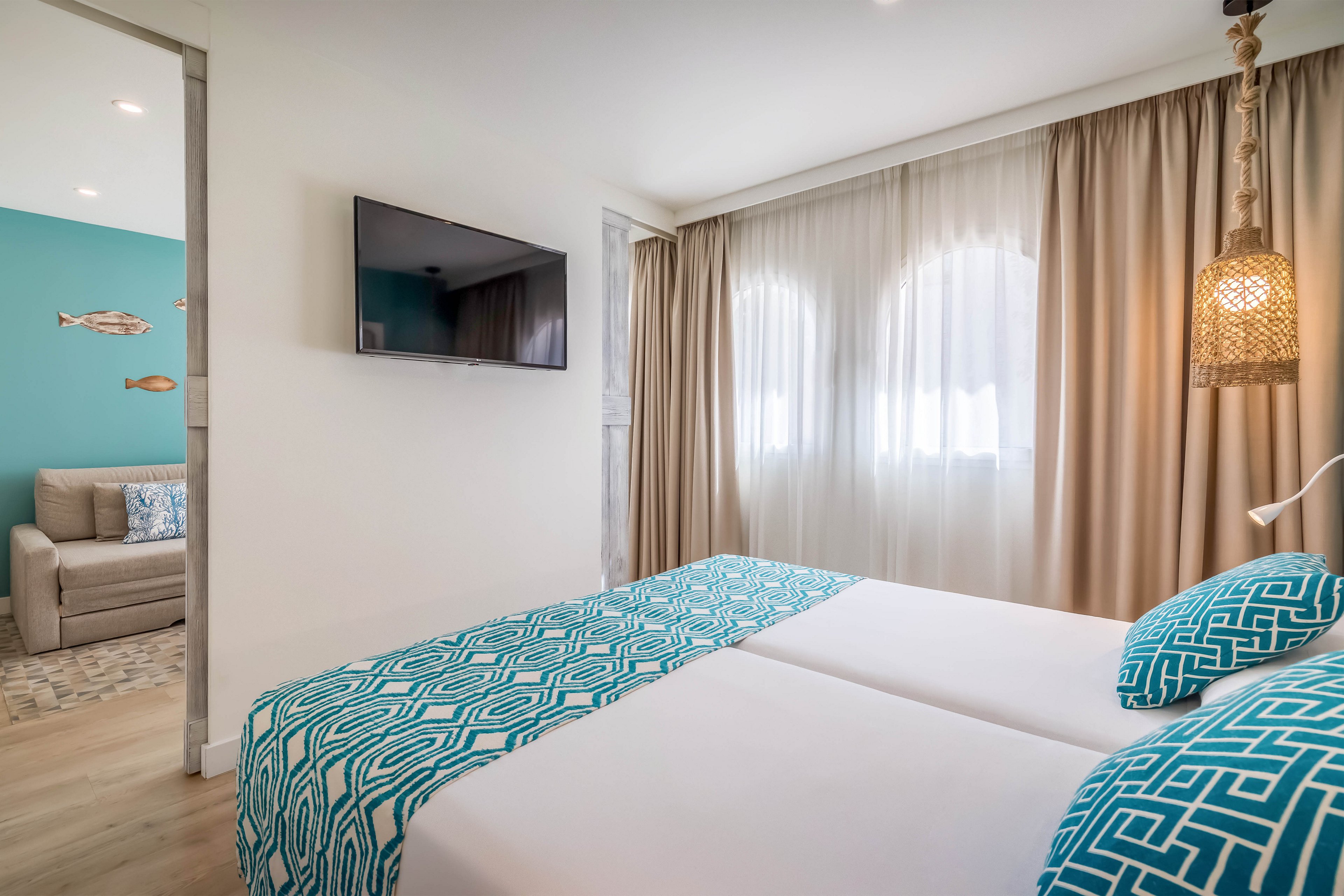 Contemporary hotel bedroom featuring two neatly made twin beds with turquoise geometric bedding. A flat-screen TV is mounted on the wall opposite the beds, and large windows with sheer and beige curtains let in soft natural light. A woven pendant lamp hangs beside the bed, adding a warm decorative touch. Through an open doorway, a cozy sitting area with a sofa and turquoise accent wall with fish decor is visible.