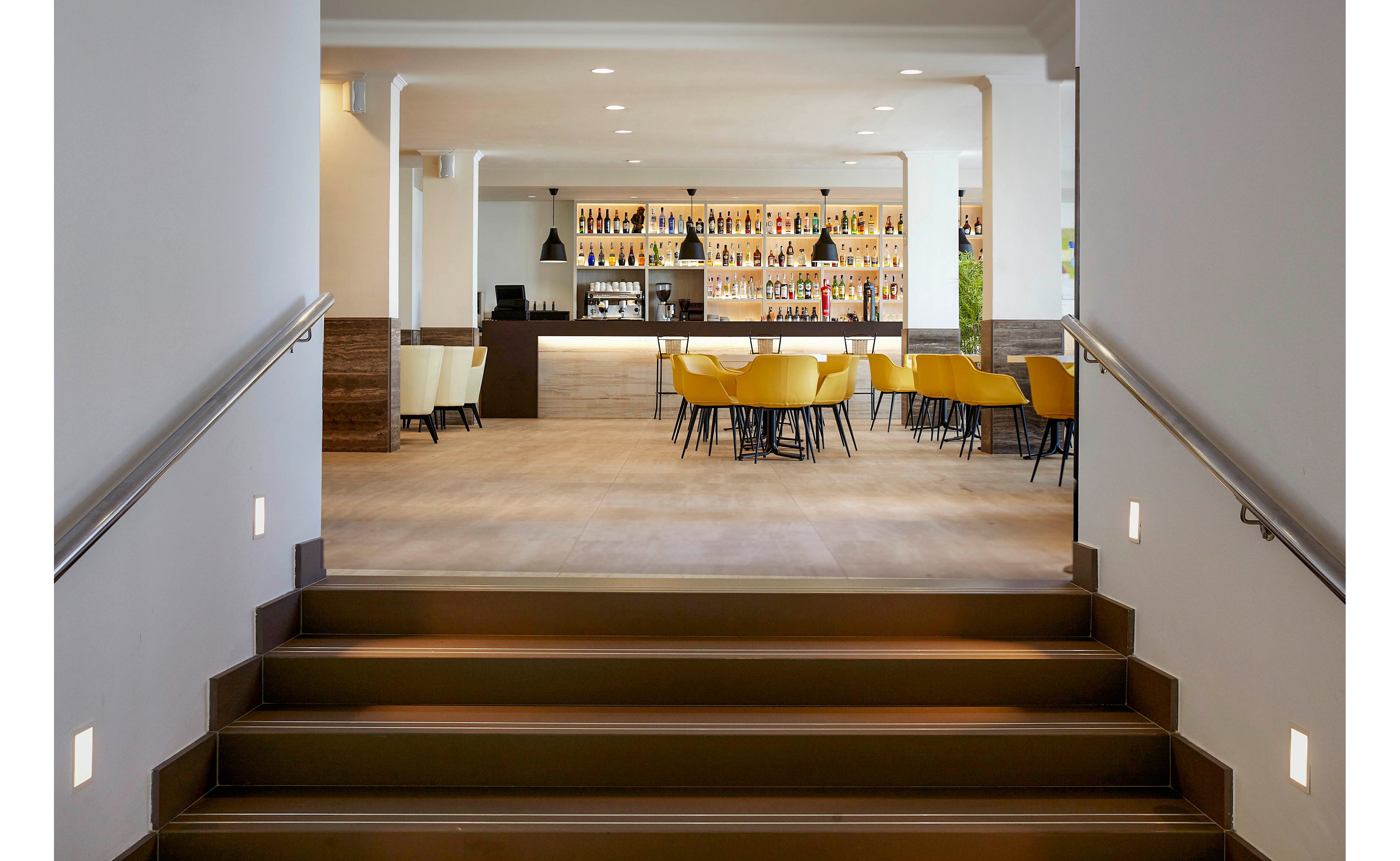 A contemporary bar area is viewed from the bottom of a short staircase with metal railings and wall lights. The open space features a long counter, shelves filled with bottles, and several yellow upholstered chairs around tables. Pendant lights hang from the ceiling, and the overall design is clean and minimal with neutral tones. The setting appears calm and inviting, suitable for socializing or relaxing.