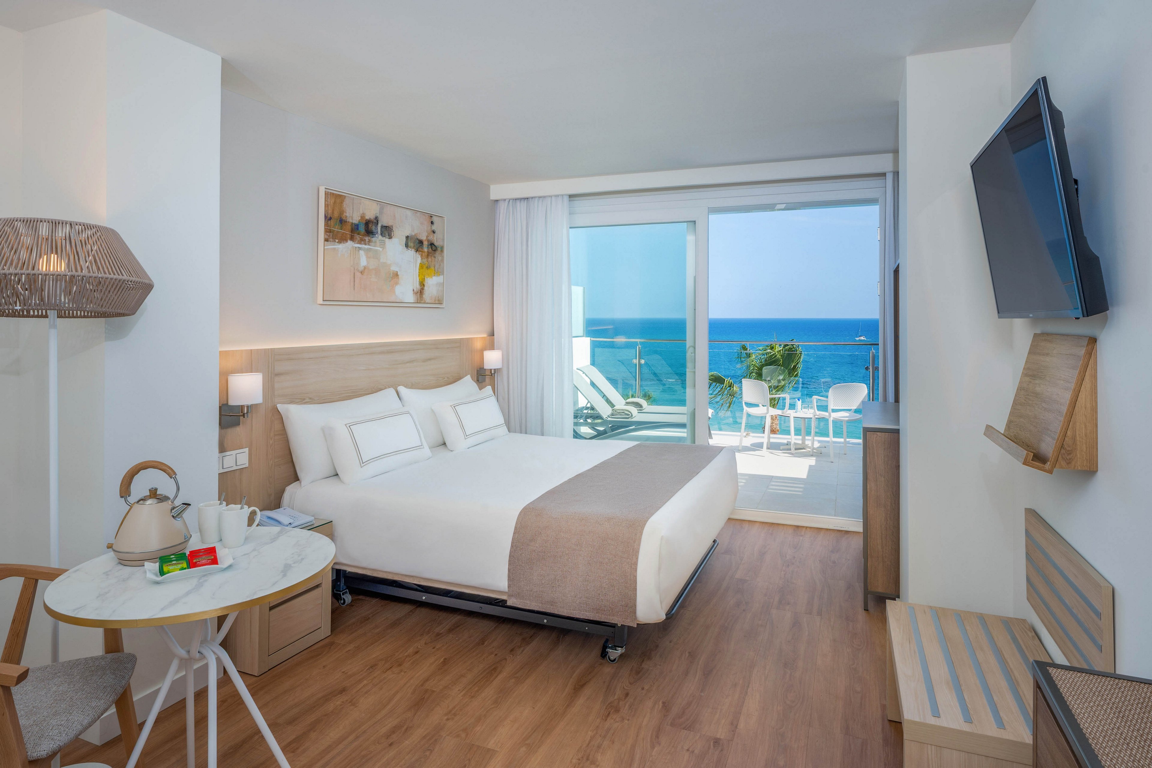 Bright contemporary hotel room featuring a neatly made double bed with white linens and beige accents. The space includes a small round table with a kettle and cups, a wall-mounted television, and light wood furniture. Large sliding glass doors open to a balcony with chairs and a clear view of the blue ocean and sky. Soft neutral tones and natural light create a calm, relaxing atmosphere ideal for a beachside stay.