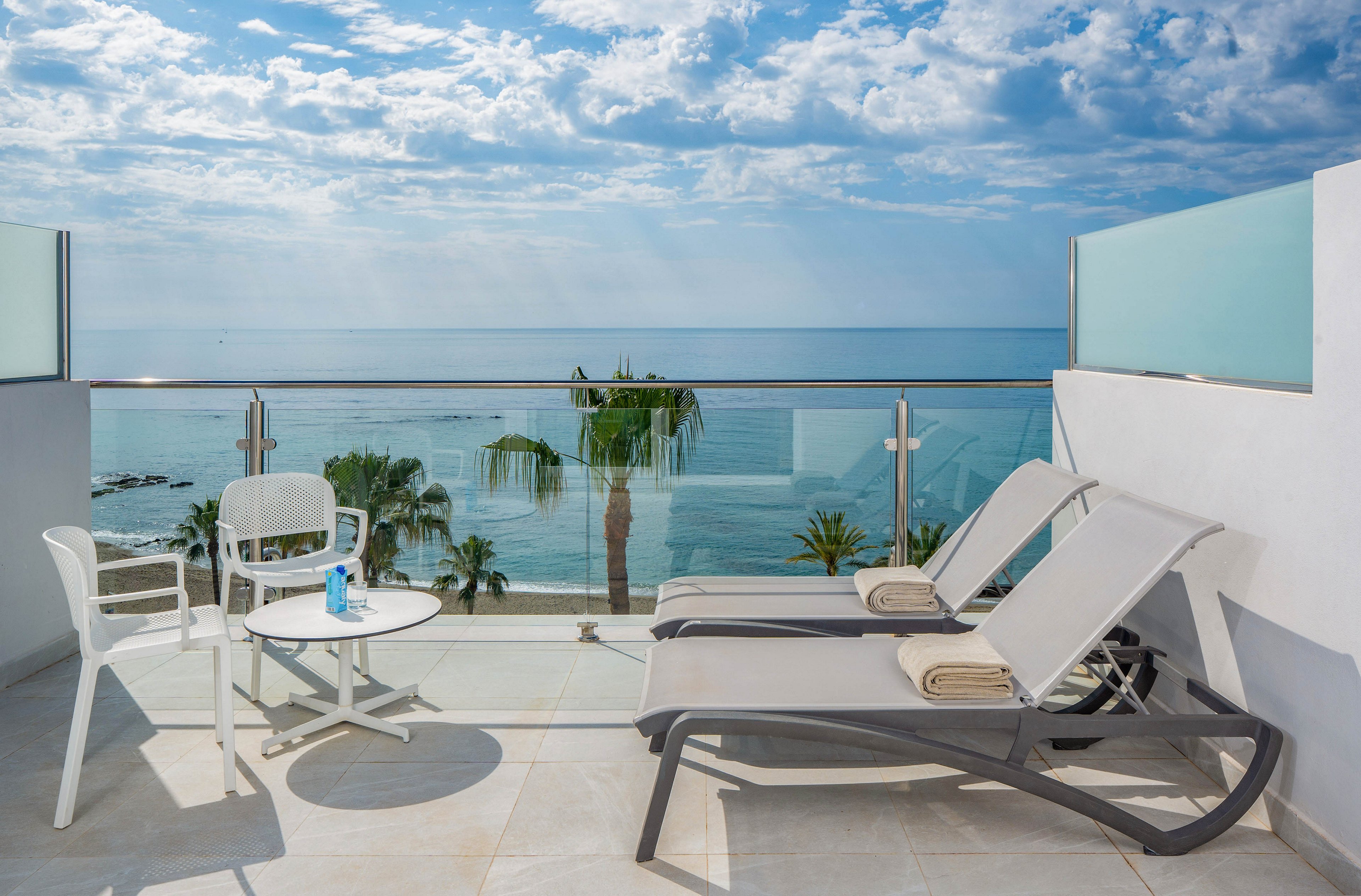 A contemporary balcony terrace features two sun loungers and a small round table with white chairs. The space overlooks a calm blue sea with palm trees and a small rocky islet in the distance. Glass railings and light-colored tiles create a clean, minimalist look under a partly cloudy sky. The scene evokes a relaxed vacation atmosphere with bright natural light and coastal views.