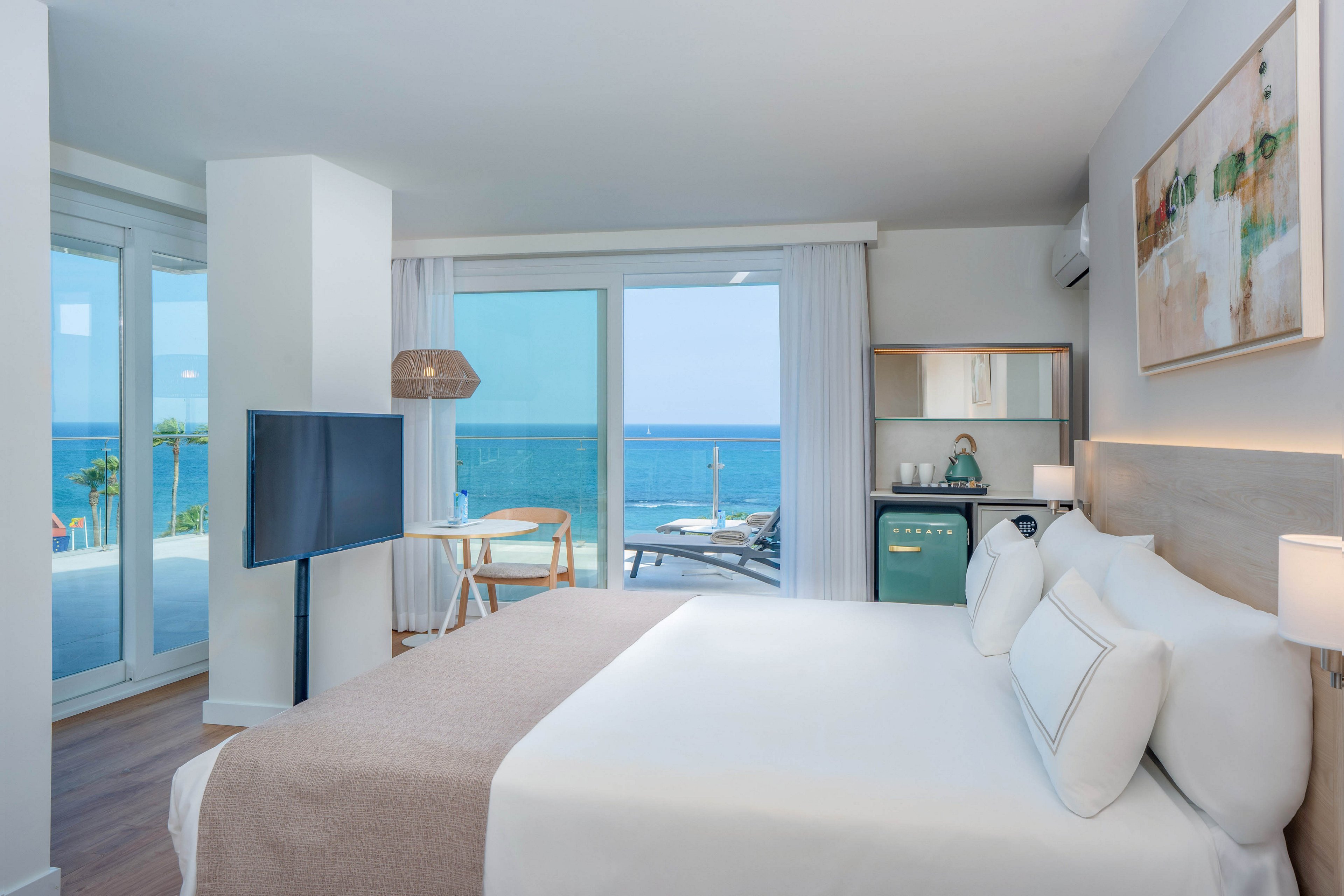 Bright contemporary hotel room features a neatly made double bed with white linens and neutral accents. Large glass doors open to a balcony overlooking a clear blue ocean and palm trees. A small dining table, flat-screen TV, and compact kitchenette with mini fridge and shelving complete the space. Soft natural light and minimalist decor create a calm, relaxing atmosphere ideal for a beach vacation.