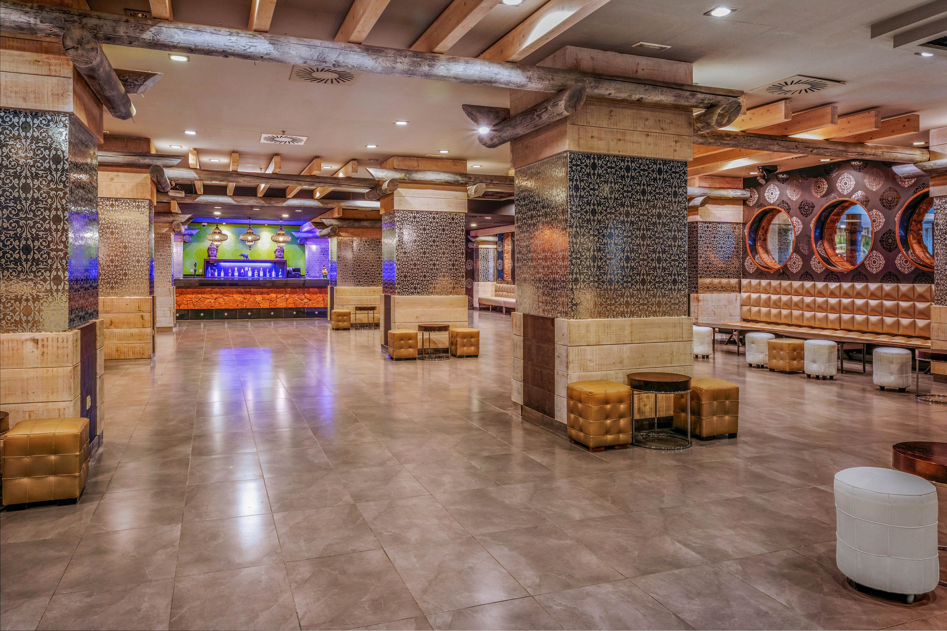 Large empty lounge interior featuring tiled columns and a polished floor. The space includes cushioned bench seating along the walls and scattered ottomans and stools. Warm lighting and wood ceiling beams create a cozy yet contemporary atmosphere. A colorful illuminated bar area is visible in the background, suggesting a venue suitable for events or nightlife.