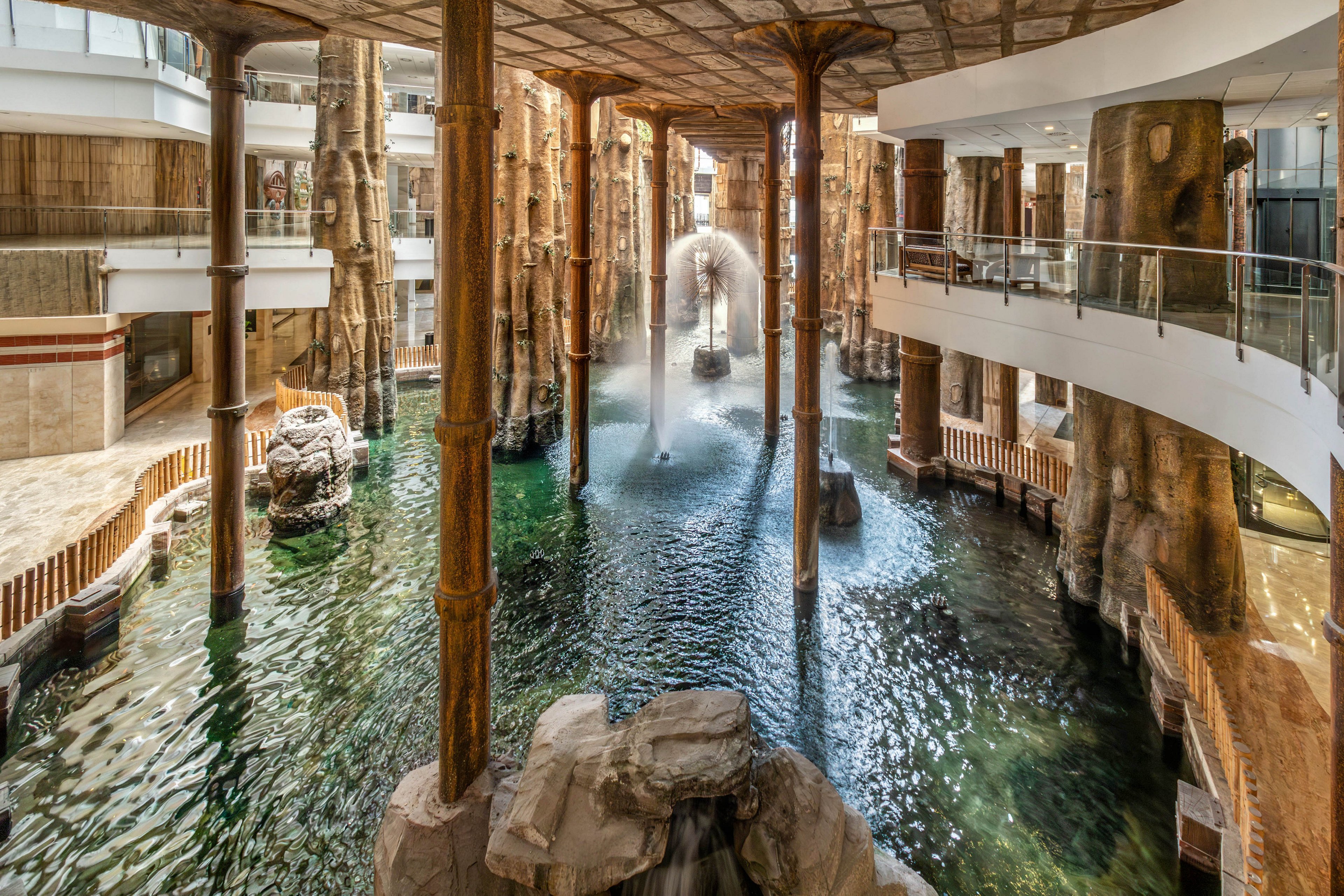 An expansive indoor water feature runs through the center of a multi-level building. Tall, tree-like pillars and rock formations rise from the water, creating a naturalistic, grotto-style atmosphere. A fountain sprays an arc of water into the air, adding movement and texture to the scene. Glass railings and walkways on the upper levels overlook the water, blending modern architecture with a resort-like interior design.