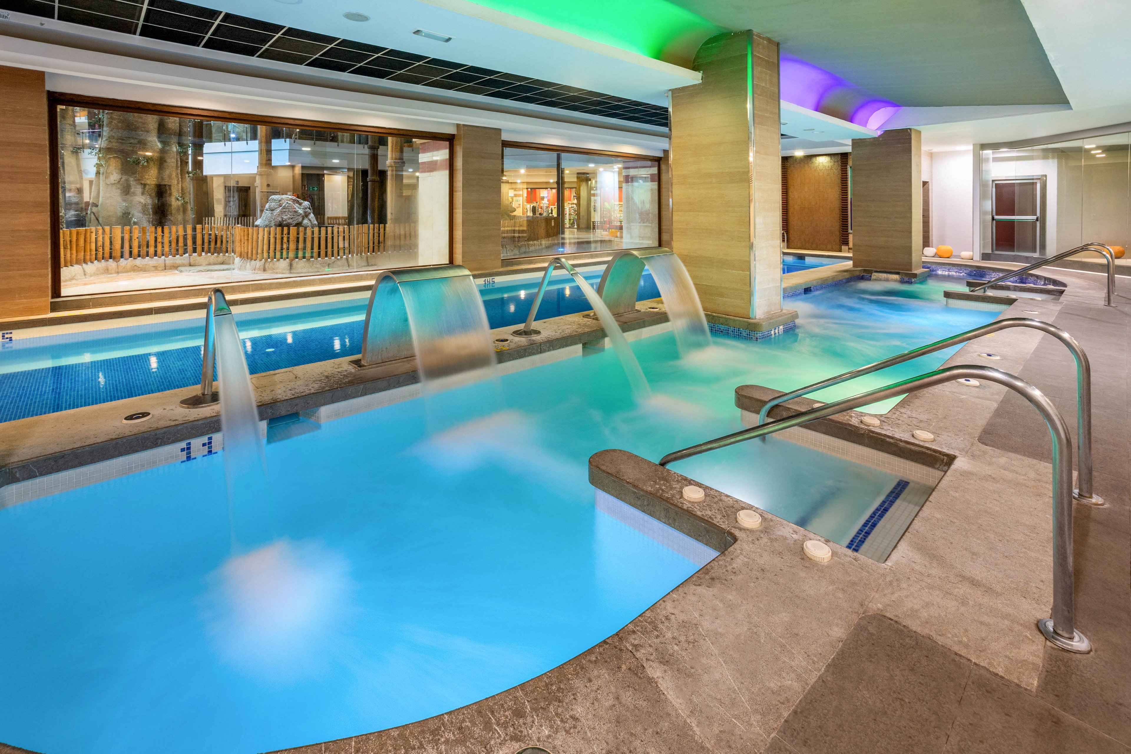 Contemporary indoor spa area featuring a long turquoise pool with powerful water jets and cascading fountains. Stainless steel handrails and steps provide safe access along the pool edge. Warm wooden wall panels and large interior windows frame the space, reflecting the water and lights. Colorful ceiling lighting adds a relaxing, luxurious atmosphere to the wellness setting.