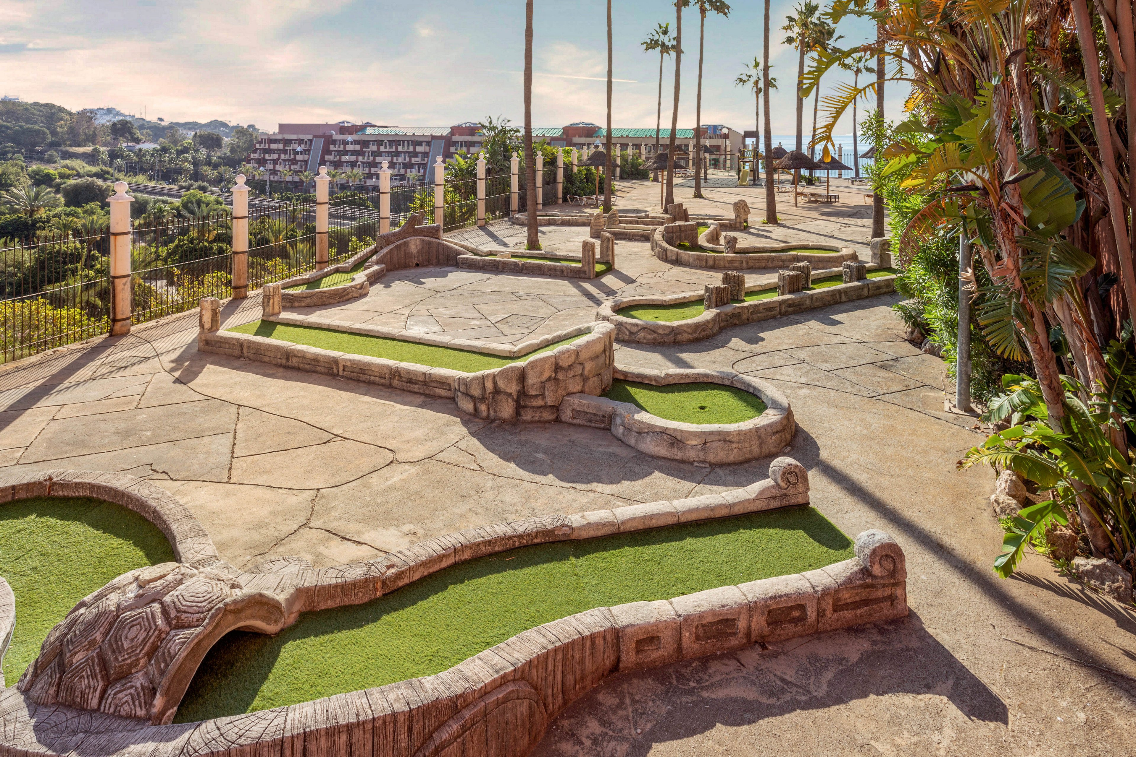 An empty outdoor mini golf course is set on a sunny hillside terrace. Curved concrete pathways and sculpted stone borders frame patches of artificial green turf. Tall palm trees and lush vegetation surround the course, with distant buildings and hills visible in the background. The warm light and clear sky create a relaxed, vacation-like atmosphere.