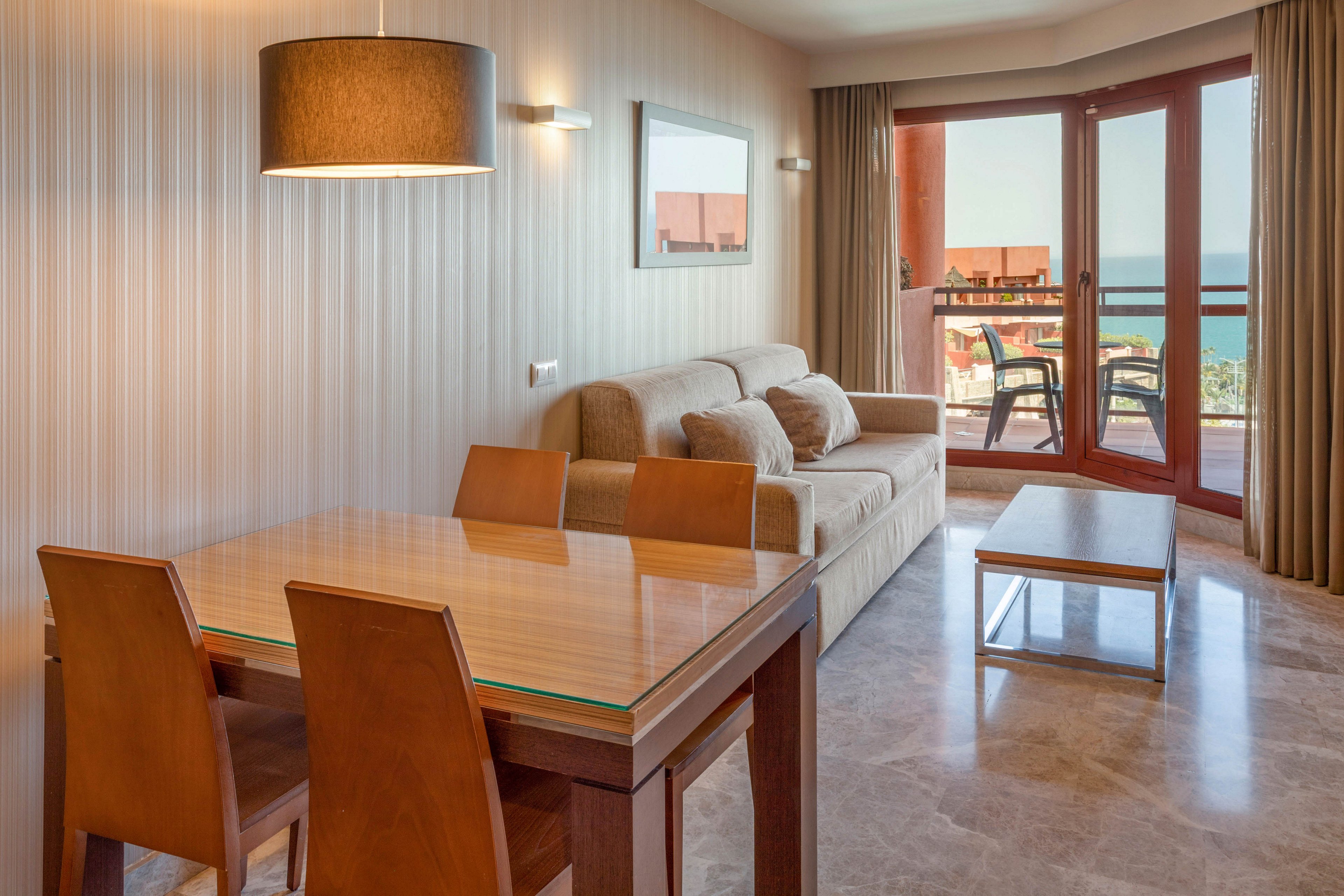 A modern hotel suite features a wooden dining table and chairs beside a comfortable beige sofa. Large floor-to-ceiling windows open onto a balcony with outdoor chairs and a view of the sea. Warm wall lights and a hanging lamp create a cozy, inviting atmosphere. Neutral tones and clean lines give the room a contemporary, minimalist style.